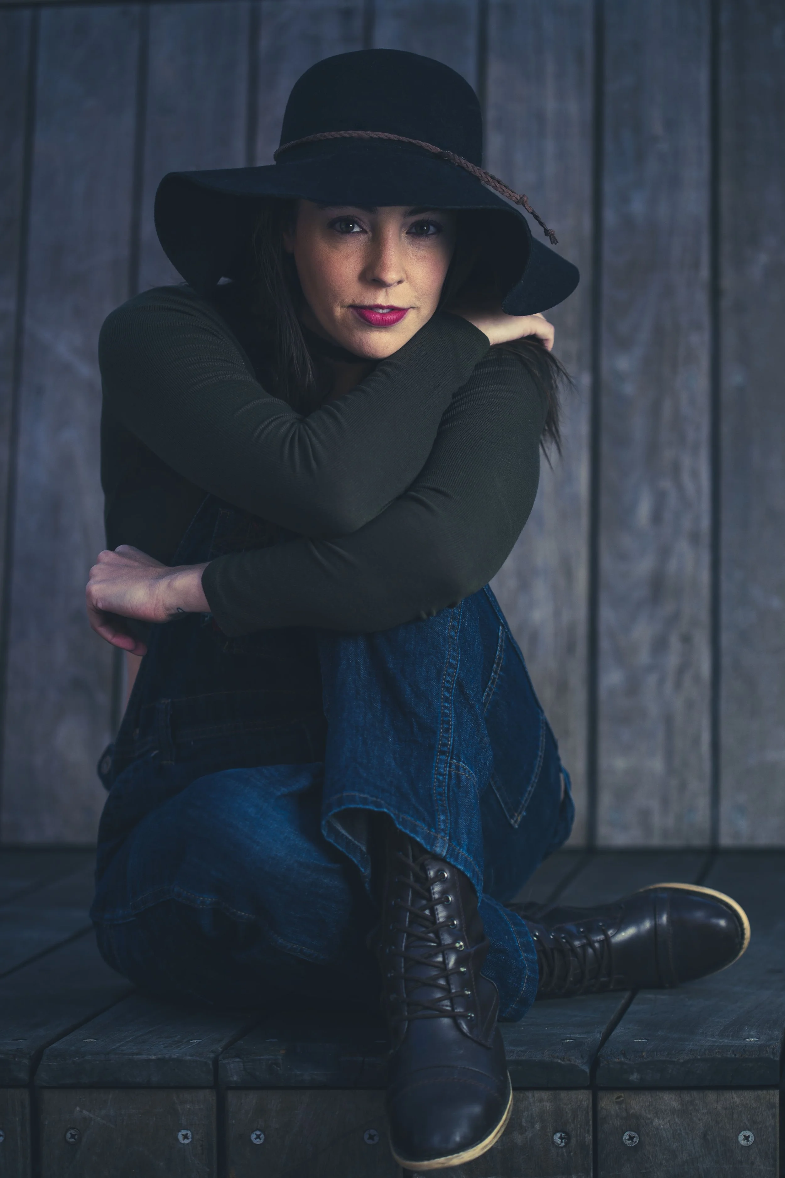 A woman sitting on a wooden surface, wearing a large black hat with a braid detail, black long-sleeve top, blue jeans, and black lace-up boots, in front of a wooden wall. BoldVision Studios