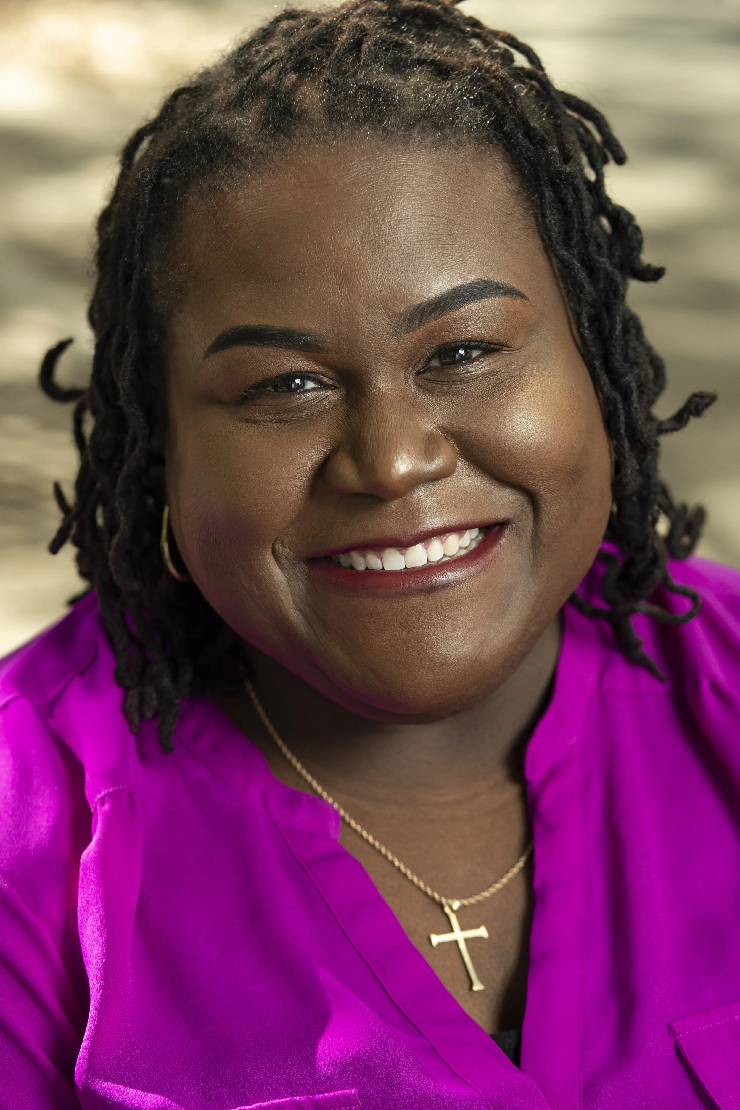 A woman with dark, styled hair smiling, wearing a bright purple top, gold cross necklace, and hoop earrings. BoldVision Studios