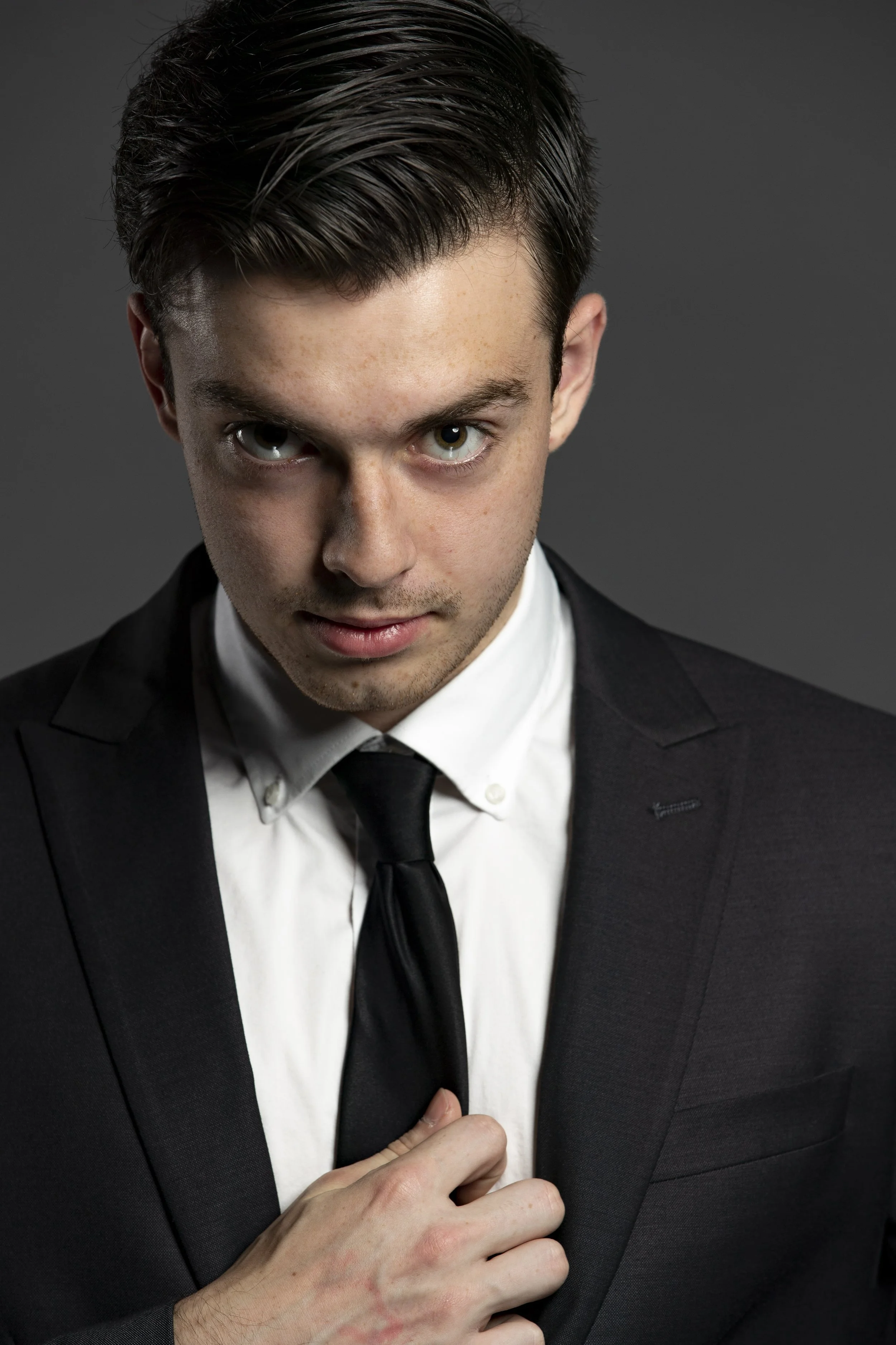 Young man in a black suit adjusting his black tie, looking directly at the camera with a confident expression, gray background. BoldVision Studios