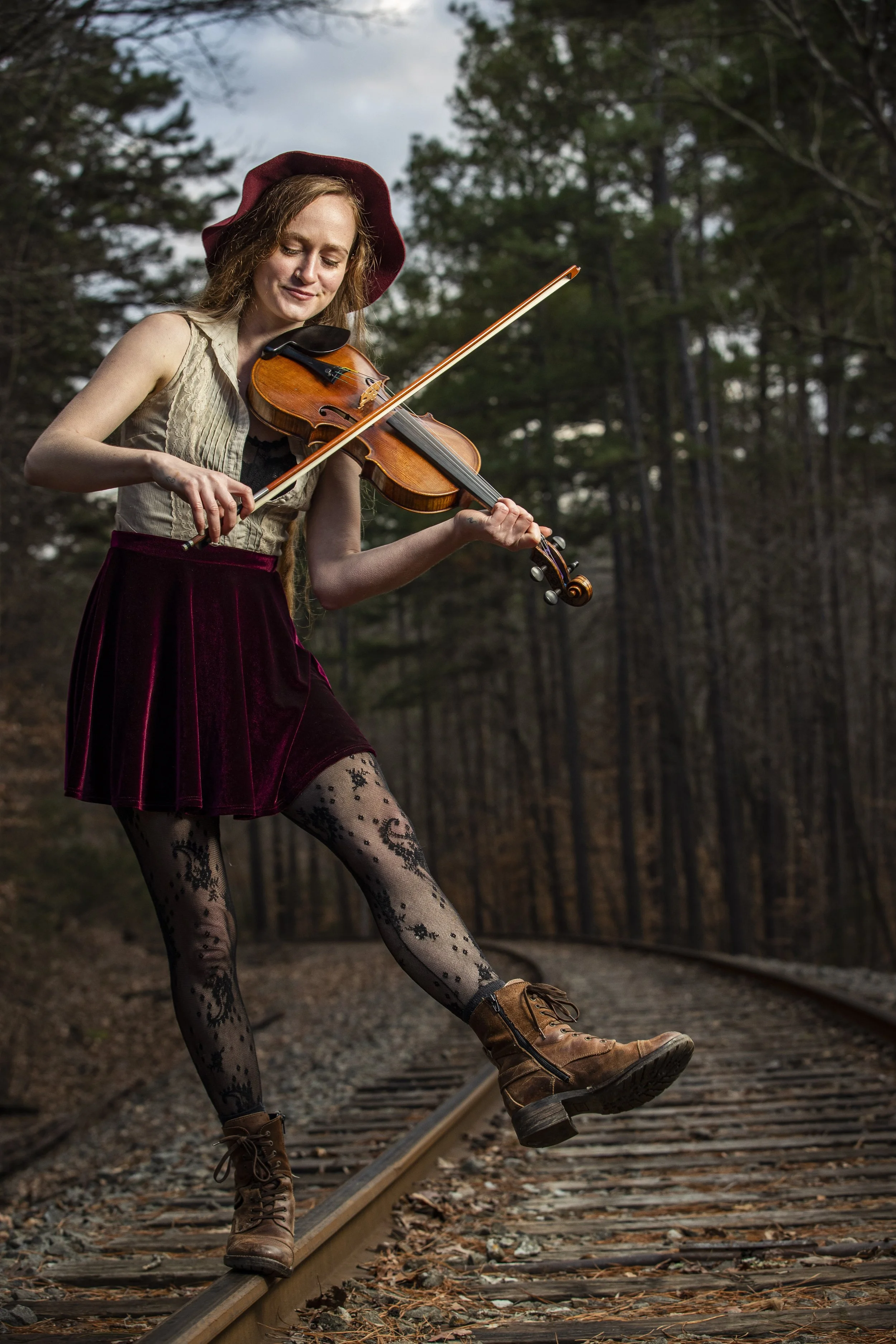 A woman wearing a red hat, a beige vest, a red skirt, patterned tights, and brown boots playing the violin by a forested railroad track. BoldVision Studios