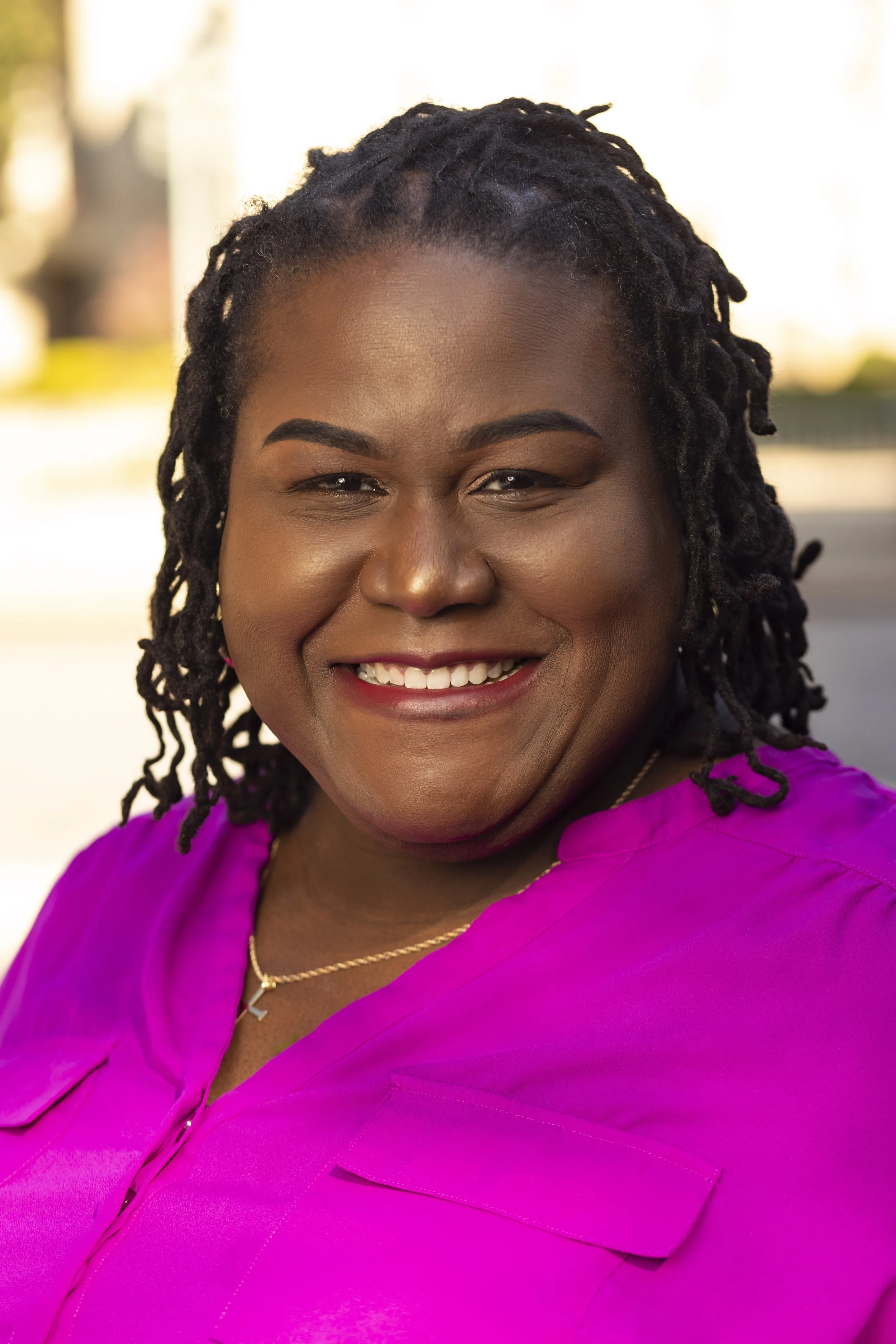 A smiling woman with dark dreadlocks, wearing a bright pink blouse and a gold necklace, outdoors with blurry background. BoldVision Studios