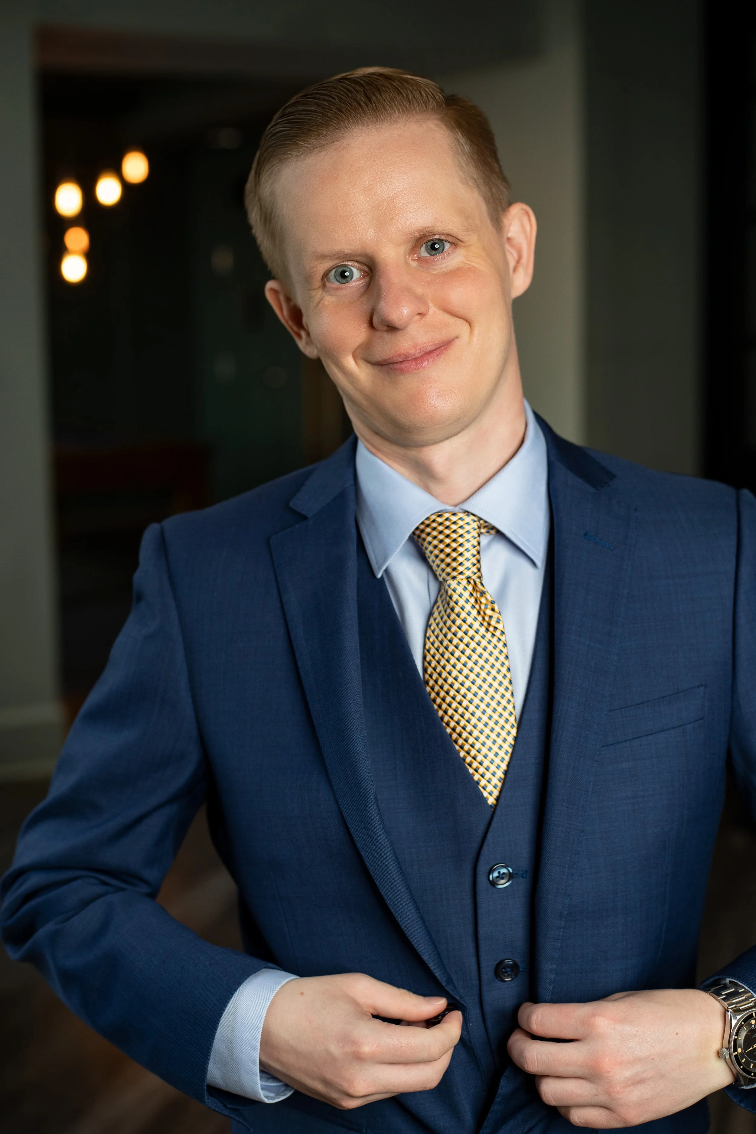 A young man with blonde hair and blue eyes wearing a blue suit, light blue shirt, and yellow patterned tie, smiling and standing indoors. BoldVision Studios