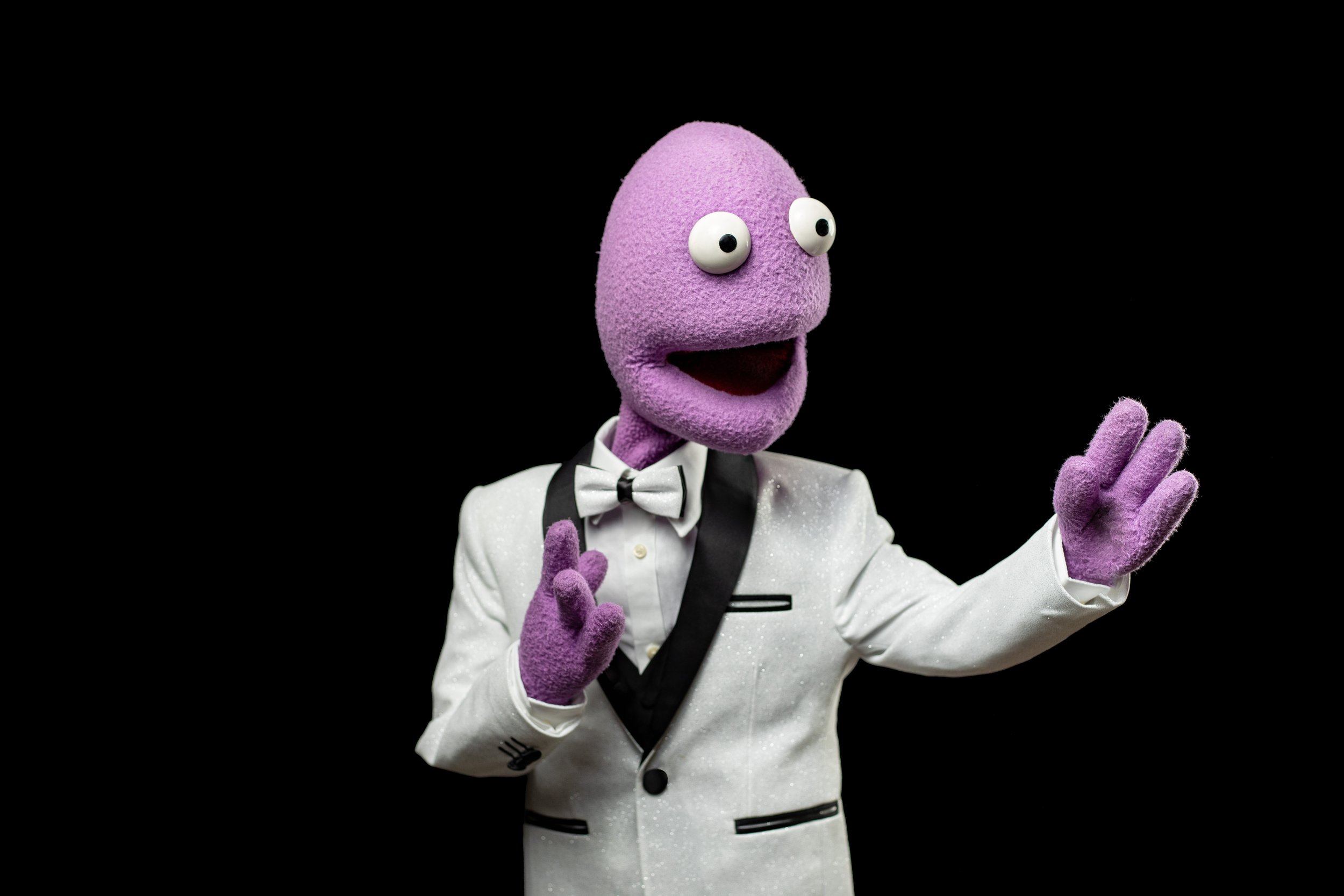 A purple puppet with big googly eyes and a big open mouth dressed in a white tuxedo and bow tie, posing against a black background. BoldVision Studios