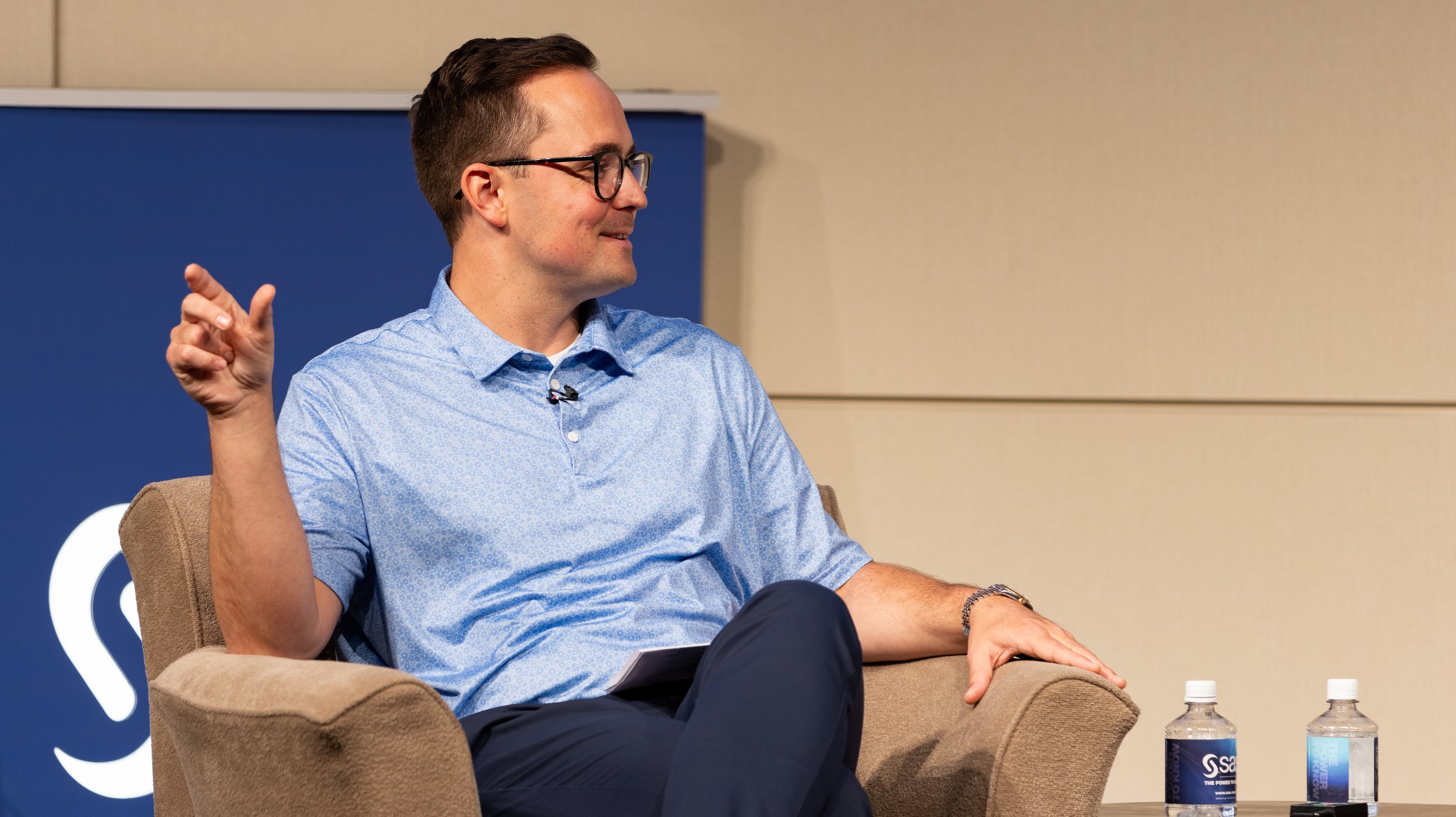 A man in glasses and a blue shirt, sitting in a beige armchair with two bottles of water on a table nearby, smiling and gesturing with his right hand during a discussion. BoldVision Studios