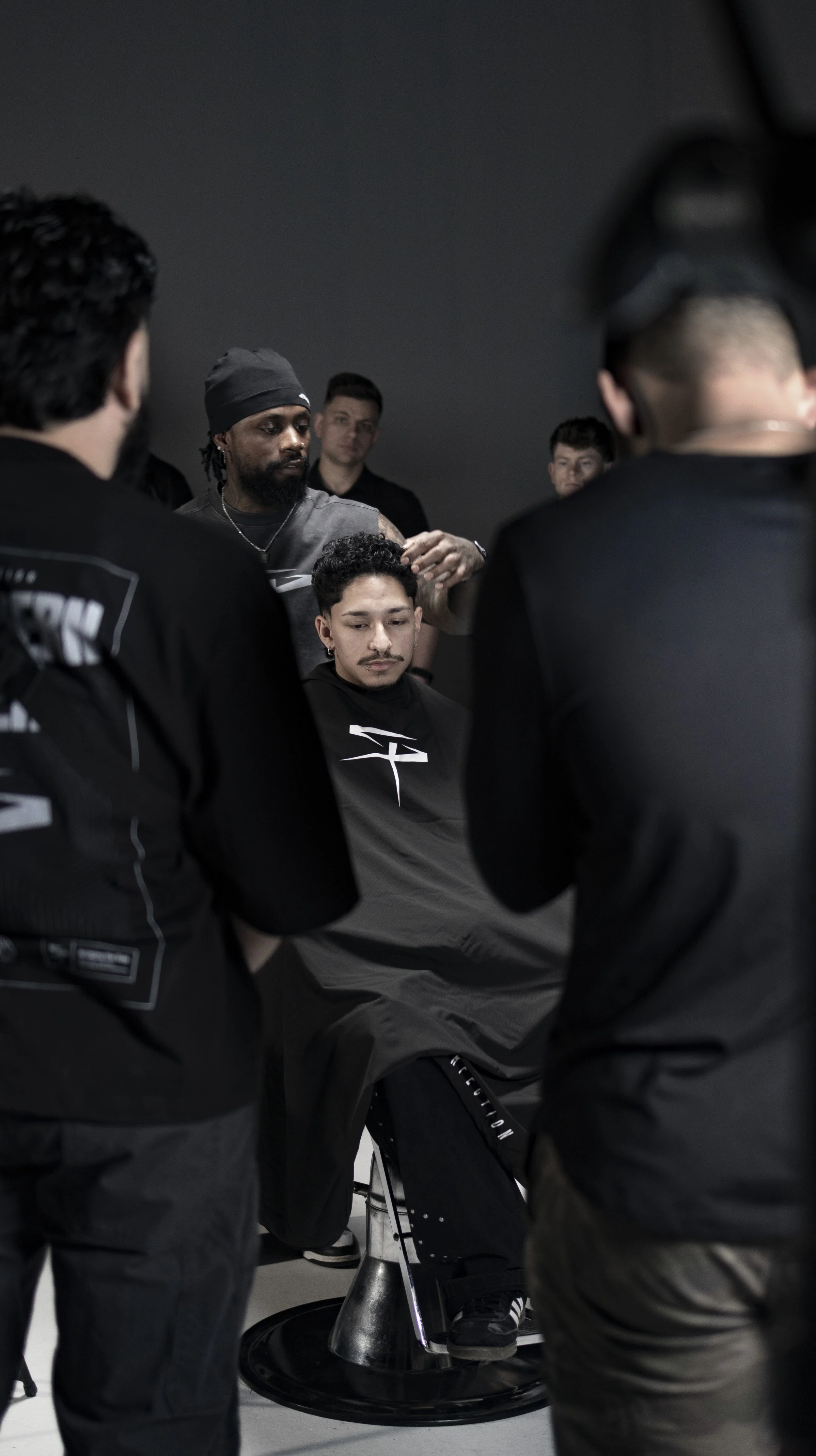 A group of young men in black clothing gathered around a seated man, who is having his hair cut or styled by a stylist in a dark indoor setting.