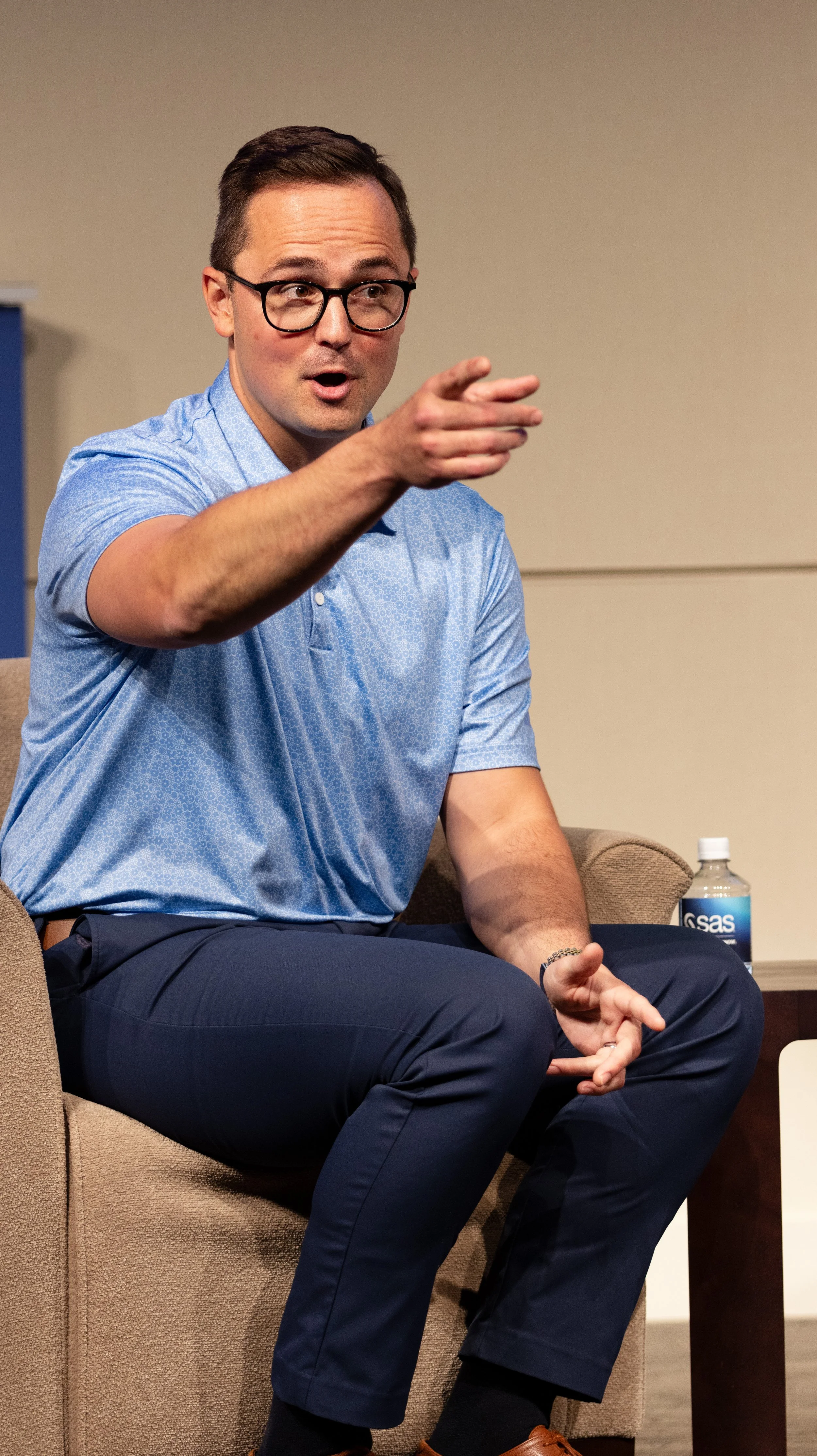 Man wearing glasses, a light blue polo shirt, and dark pants sitting on a beige chair, gesturing with his right hand during a discussion or presentation. BoldVision Studios