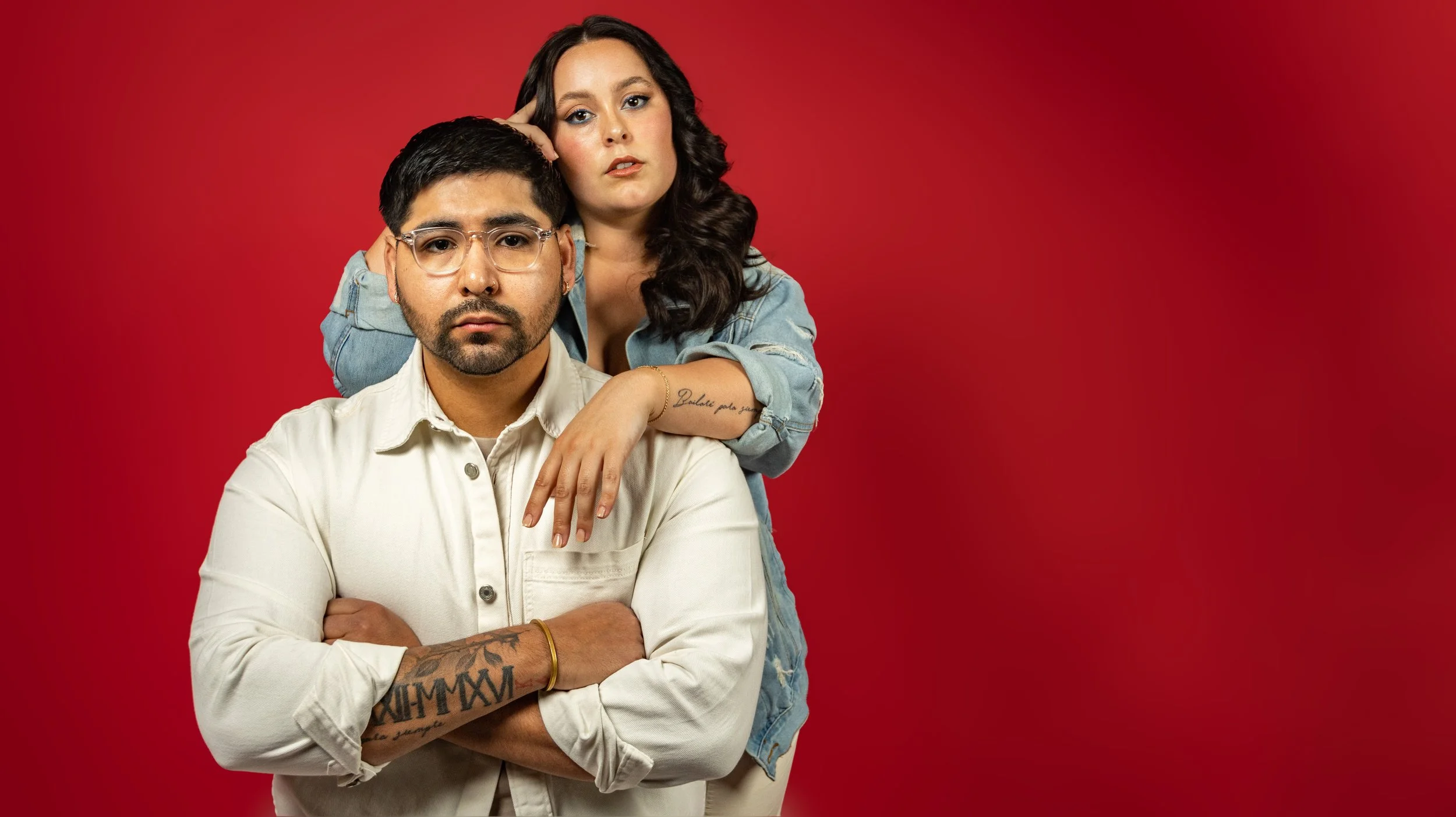 A man in glasses with a beard and tattoos on his arm stands with his arms crossed, a woman with long dark hair leaning on his shoulder, both against a red background. BoldVision Studios