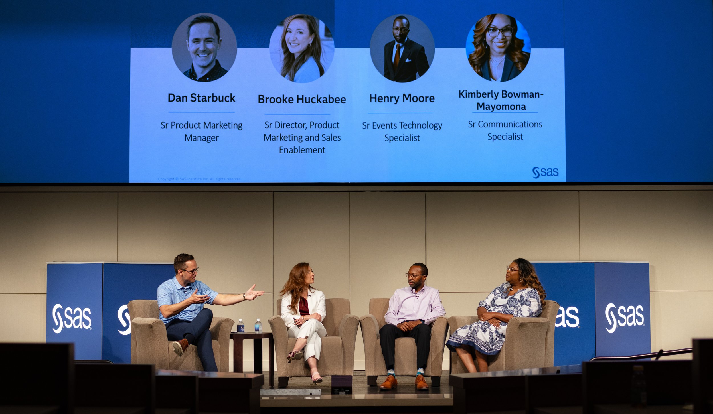Panel discussion at SAS conference featuring four speakers seated on stage with a large screen displaying their names and titles behind them. BoldVision Studios
