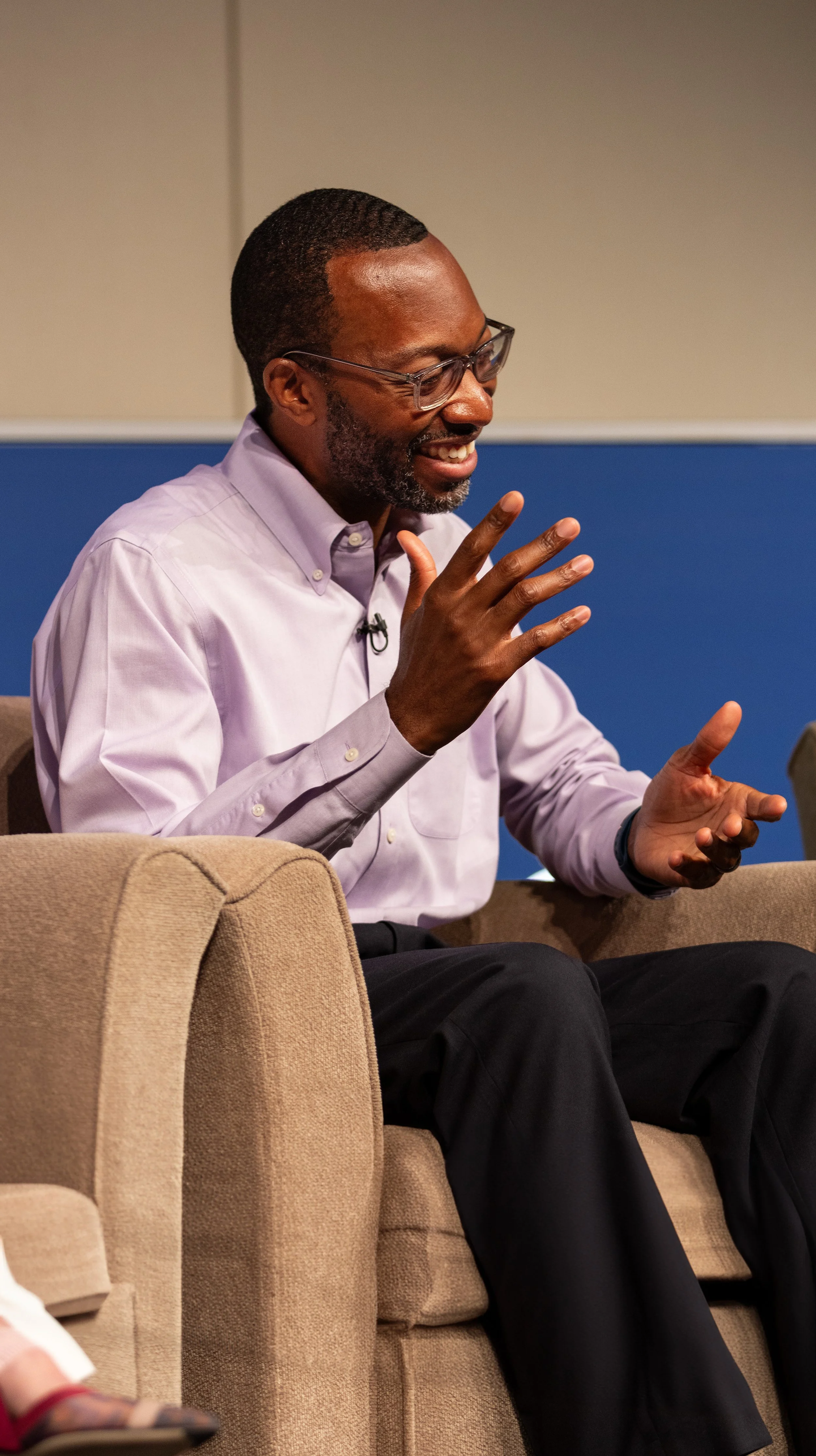 A man with glasses, a beard, and a mustache, wearing a purple dress shirt, smiling and talking while sitting on a beige sofa. BoldVision Studios