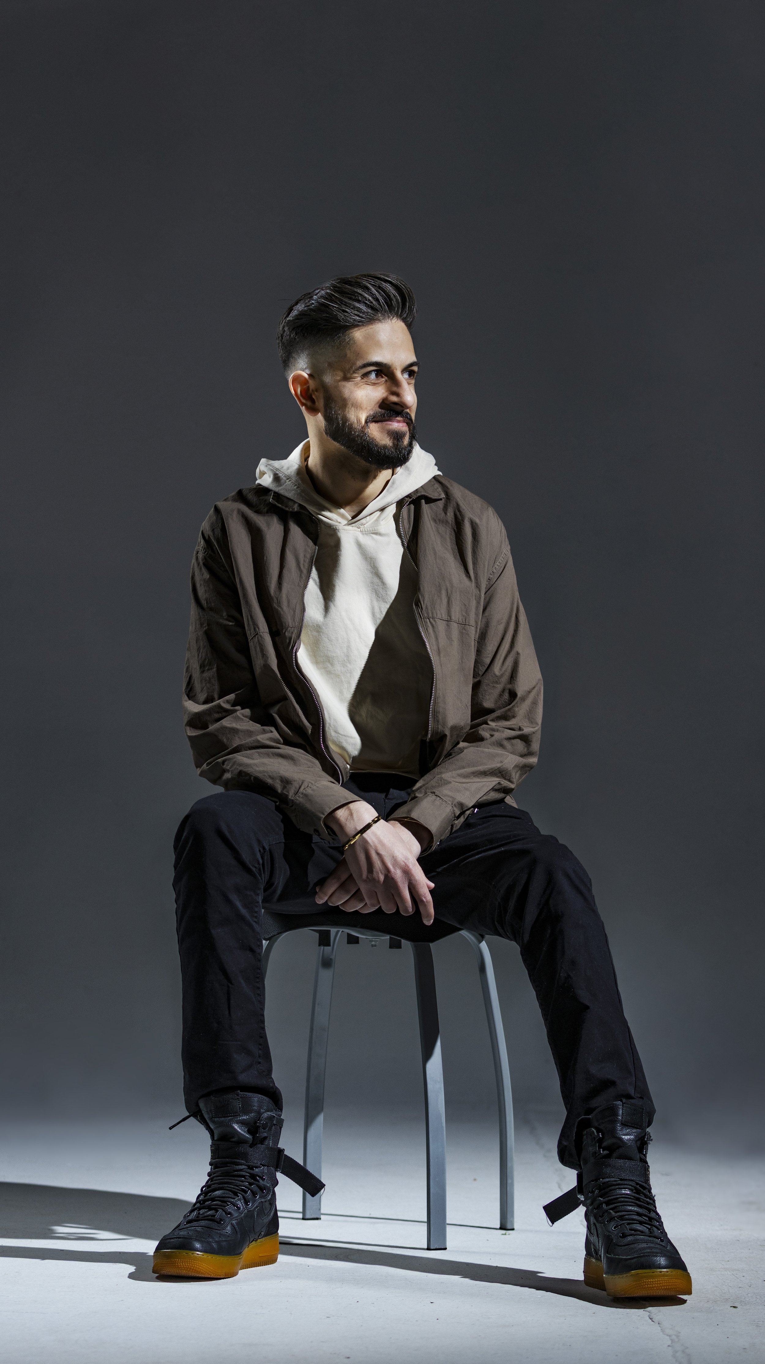 A young man with a beard and styled hair sitting on a chair against a plain, dark background, wearing a brown jacket, a white hoodie, black pants, and black boots with yellow soles. BoldVision Studios