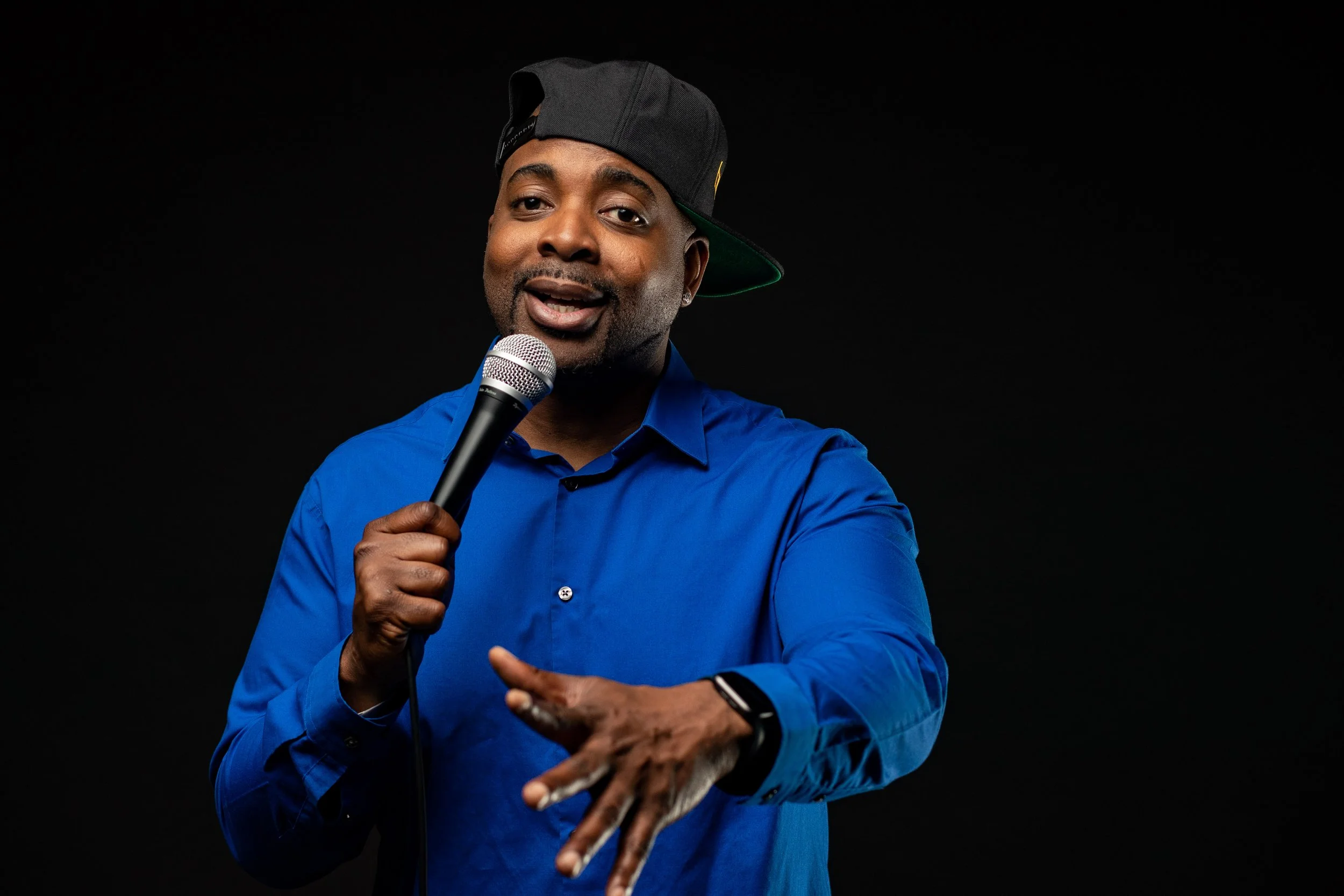 A man wearing a blue shirt, black baseball cap tilted sideways, holding a microphone and performing on stage against a black background. BoldVision Studios