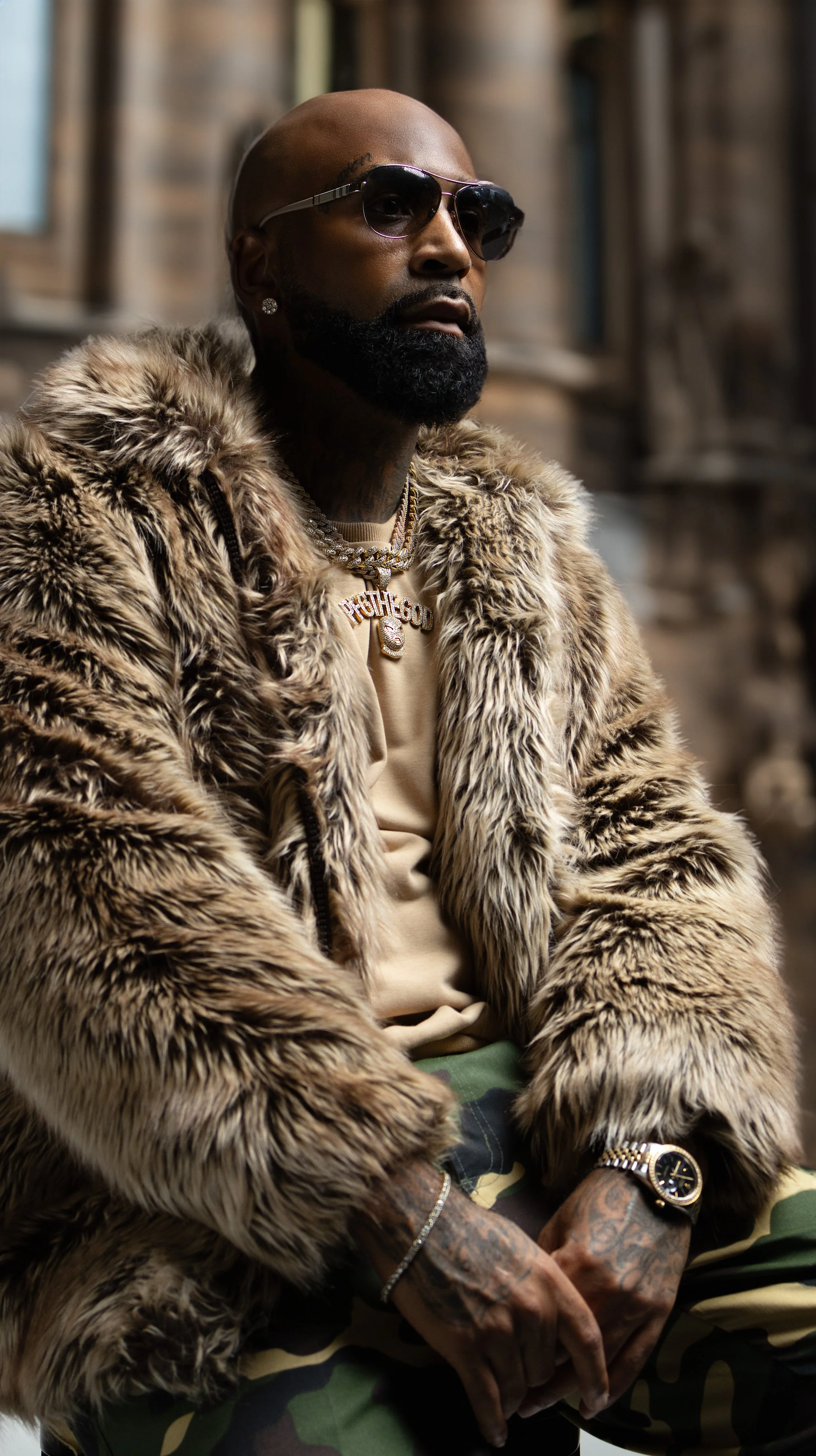 A man with a beard and sunglasses wearing a fur coat, gold jewelry, and camouflage pants, sitting outdoors. BoldVision Studios