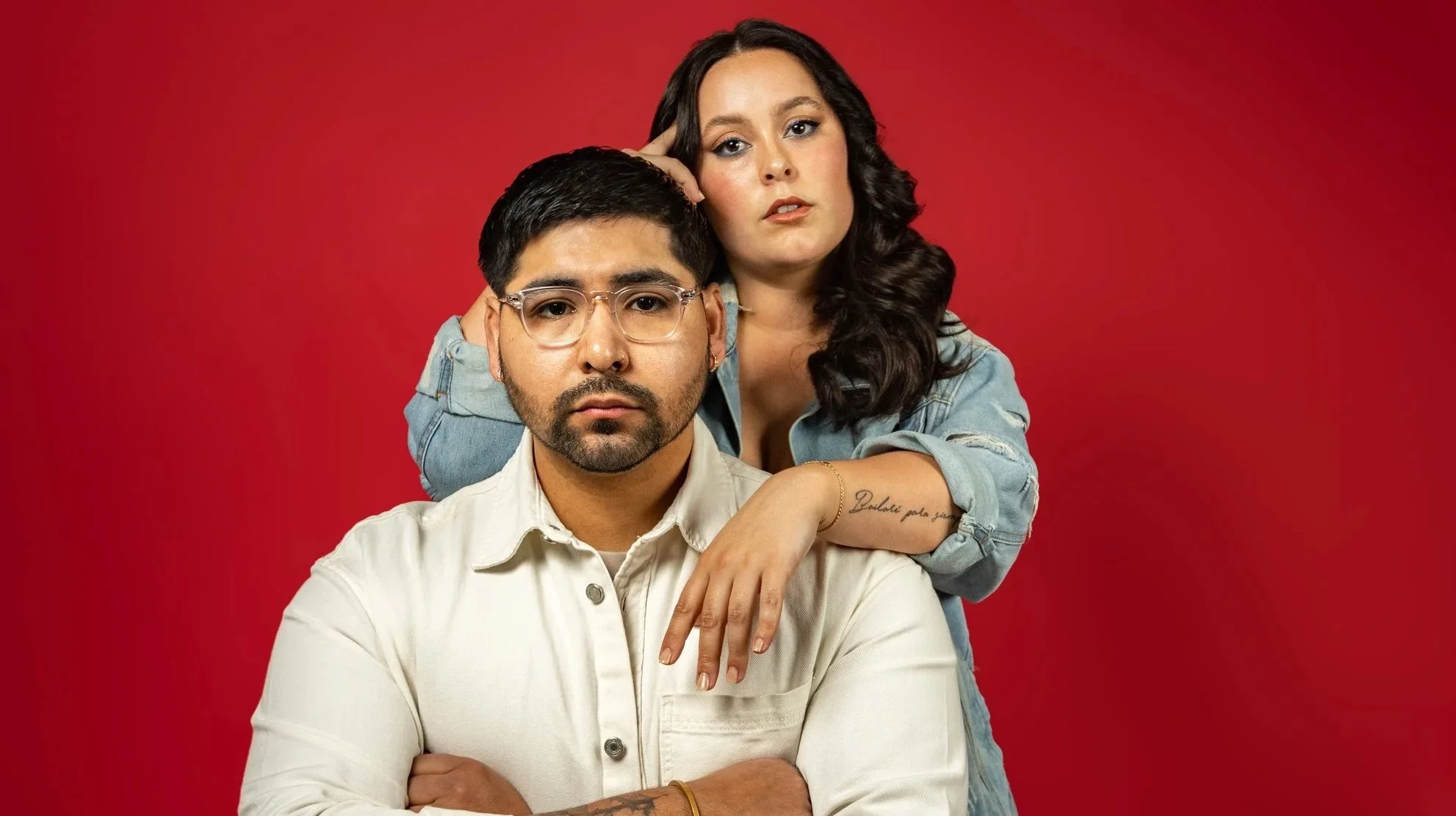 Two people, a man sitting with arms crossed and a woman standing behind him with her arm resting on his shoulder against a red background.