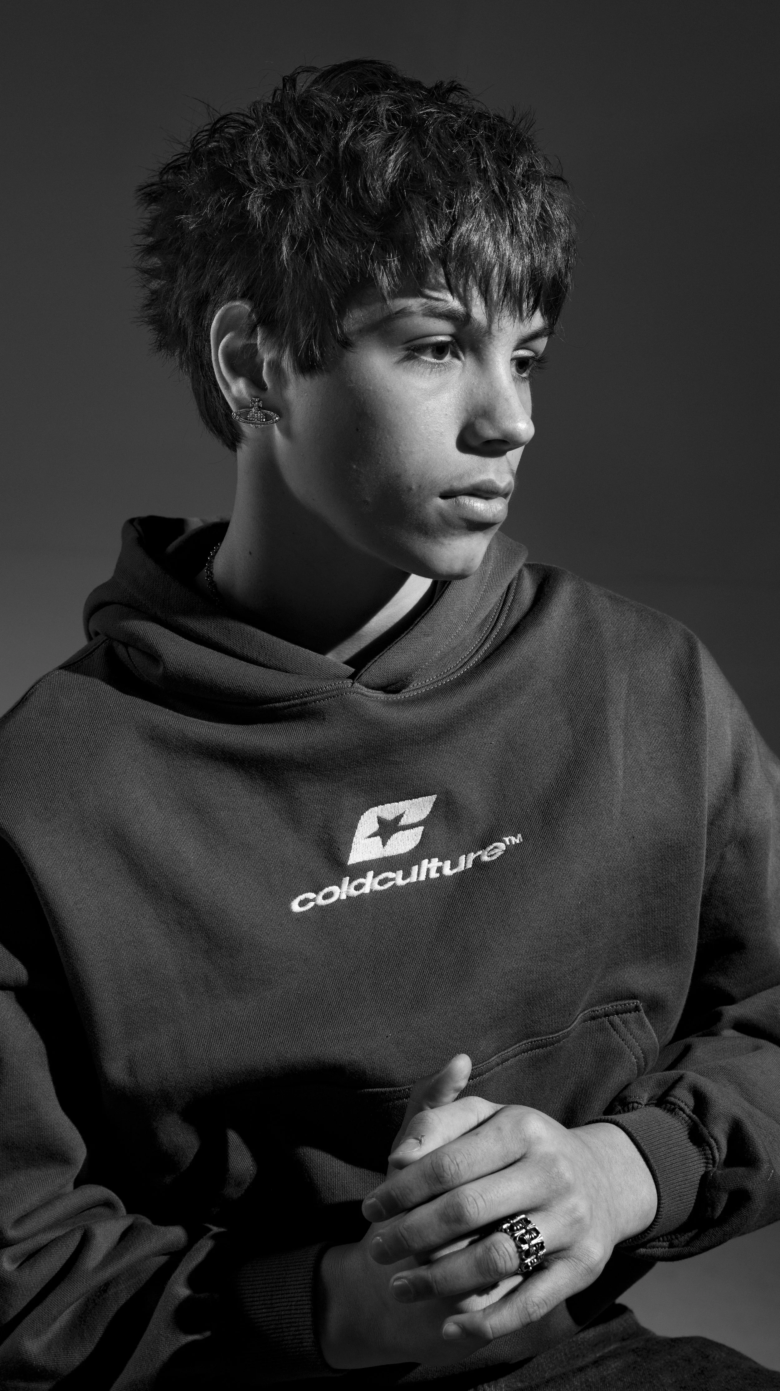 Black and white portrait of a young person with short, textured hair wearing a hoodie with Cold Culture logo, looking to the side with hands clasped. BoldVision Studios