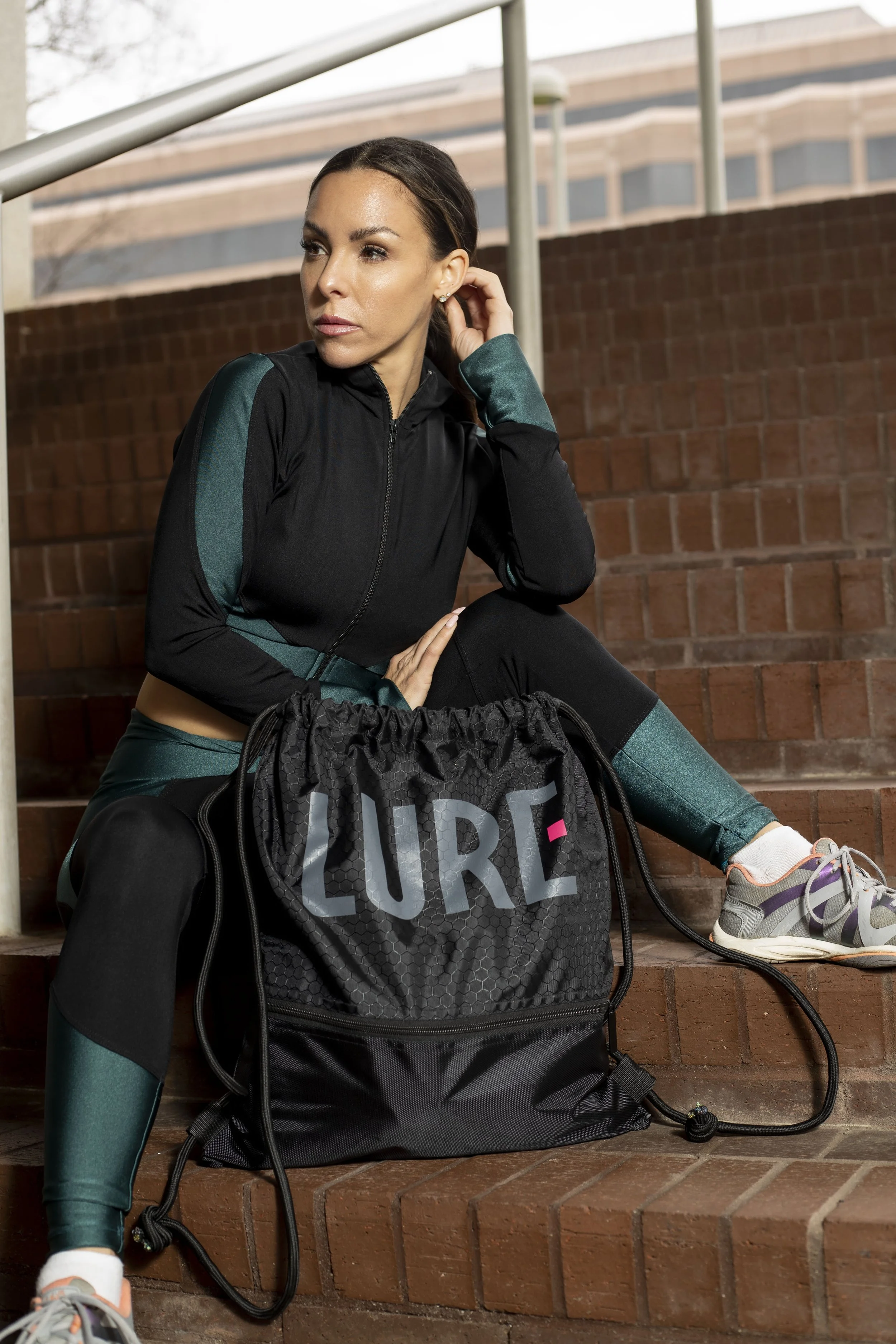 A woman in athletic wear sitting on brick stairs outdoors next to a black workout bag with the word 'LURE' on it. BoldVision Studios