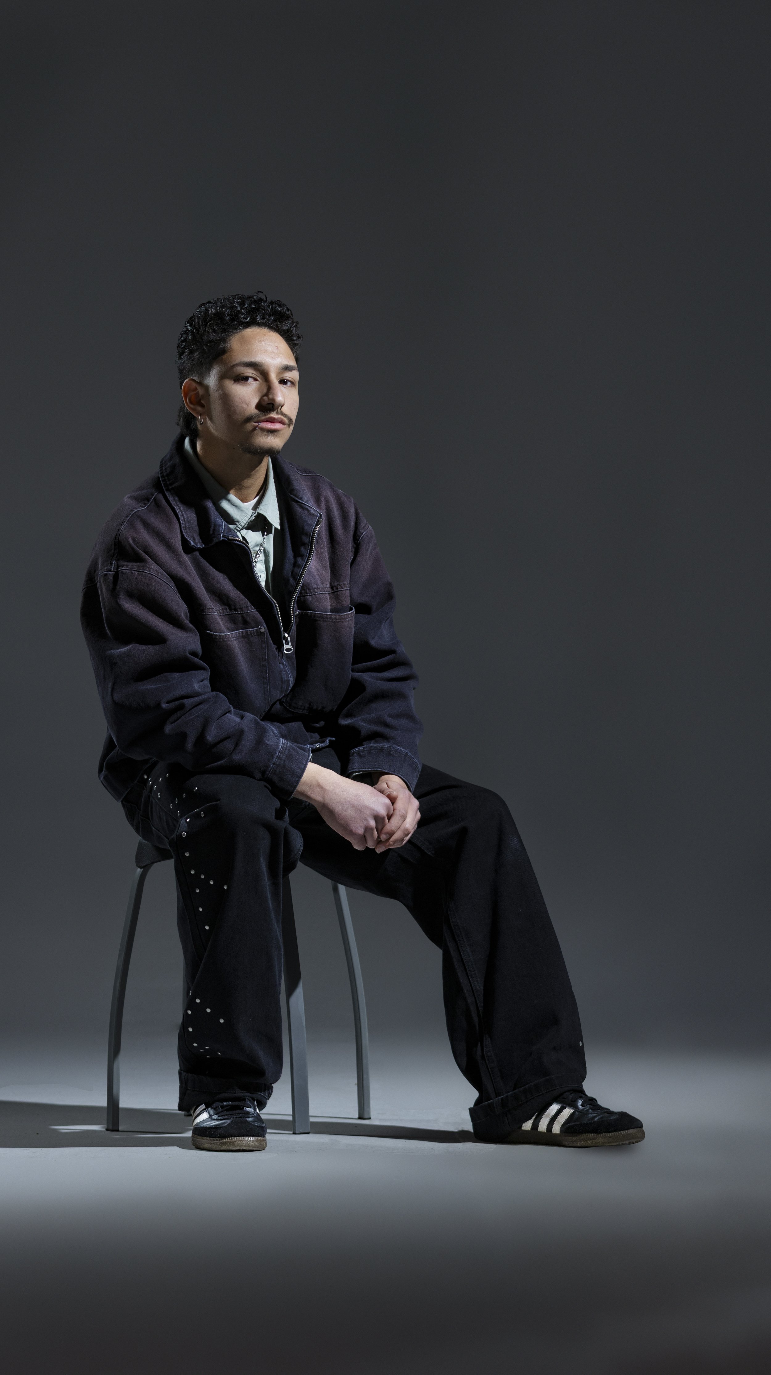 A young man is sitting on a stool against a plain, dark background. He is wearing a dark jacket, dark pants with silver studs, and black sneakers with white stripes. He has short, curly black hair and a light mustache, with a neutral expression on hi