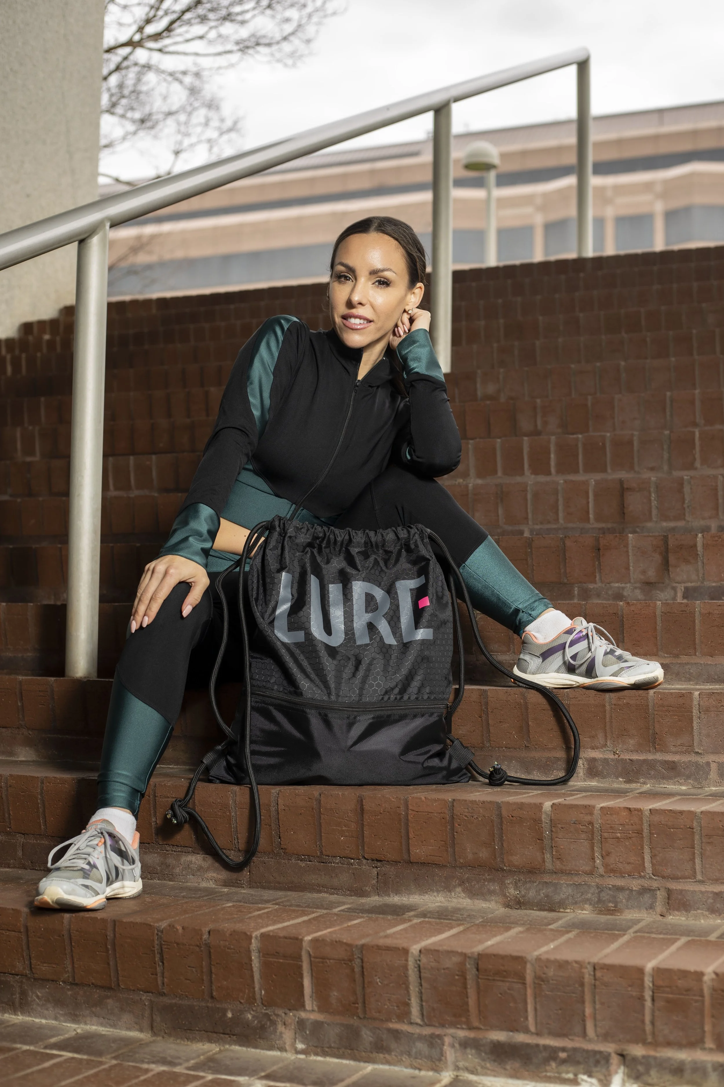 A woman in workout clothes sitting on brick stairs with a large black bag labeled 'LURE' next to her, outdoors, with a building and trees in the background. BoldVision Studios
