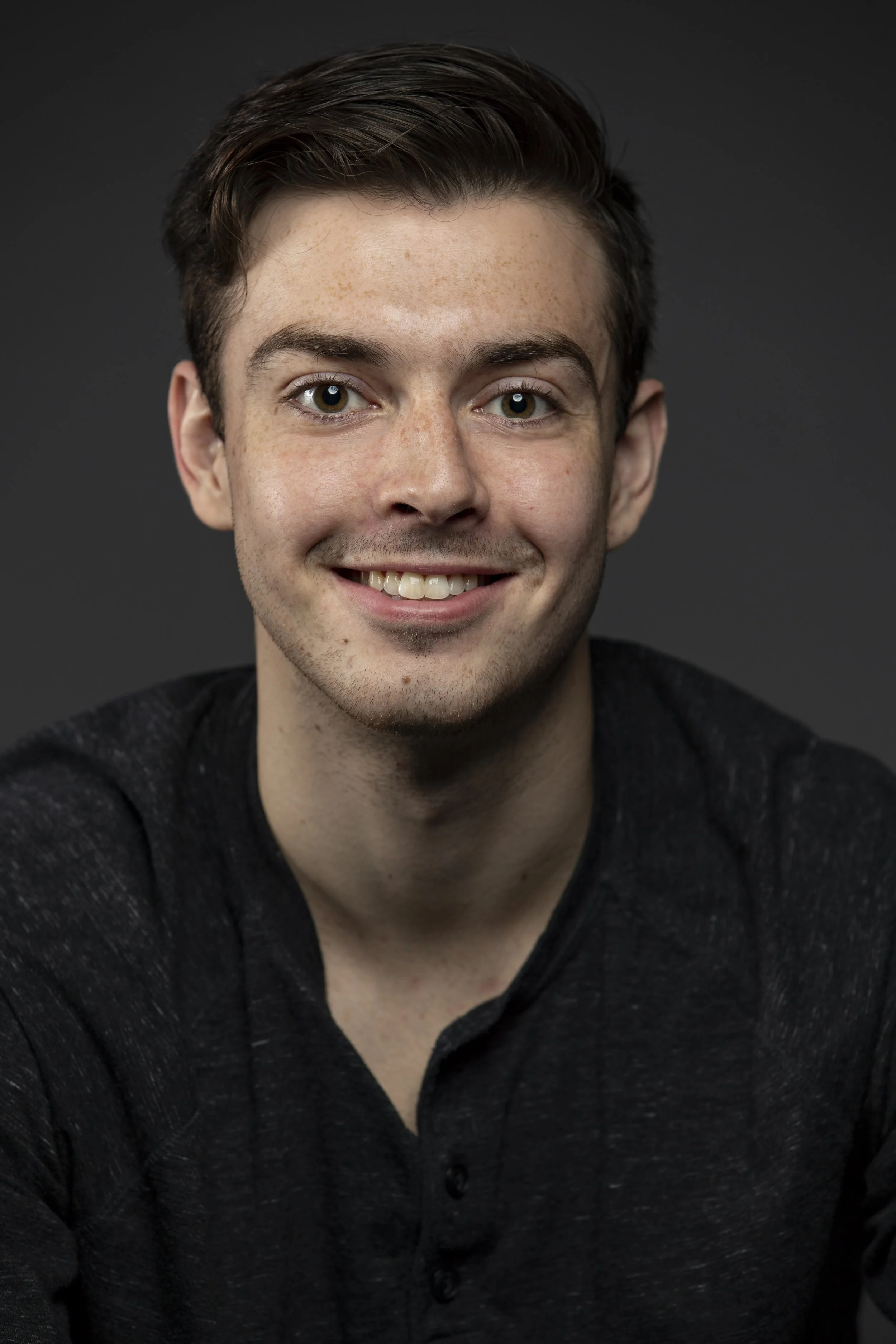 Portrait of a young man with short brown hair, light skin, and green eyes, smiling, against a dark background. BoldVision Studios