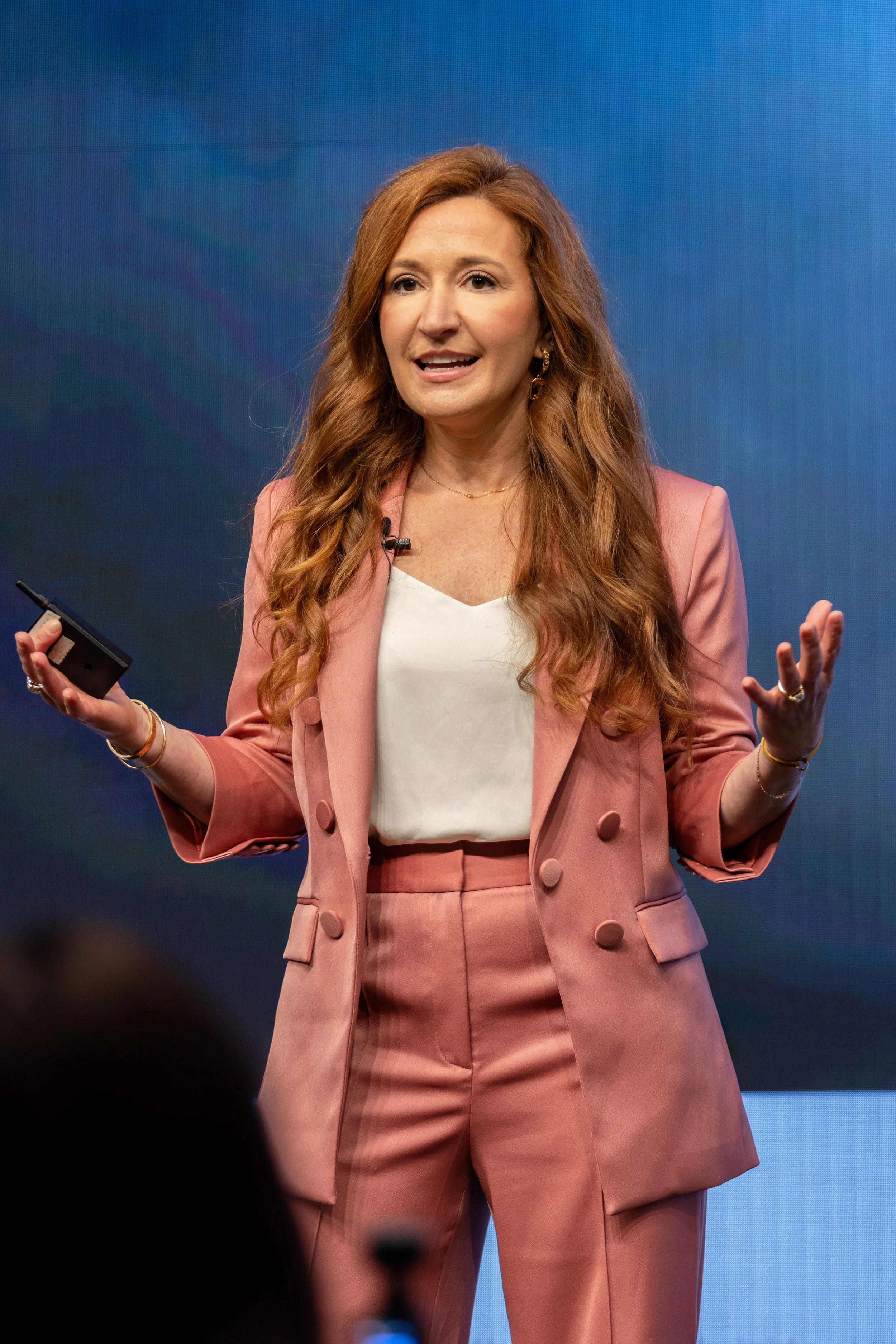 A woman with long red hair, in a pink suit, gesturing with her hands while speaking in front of a blue background.