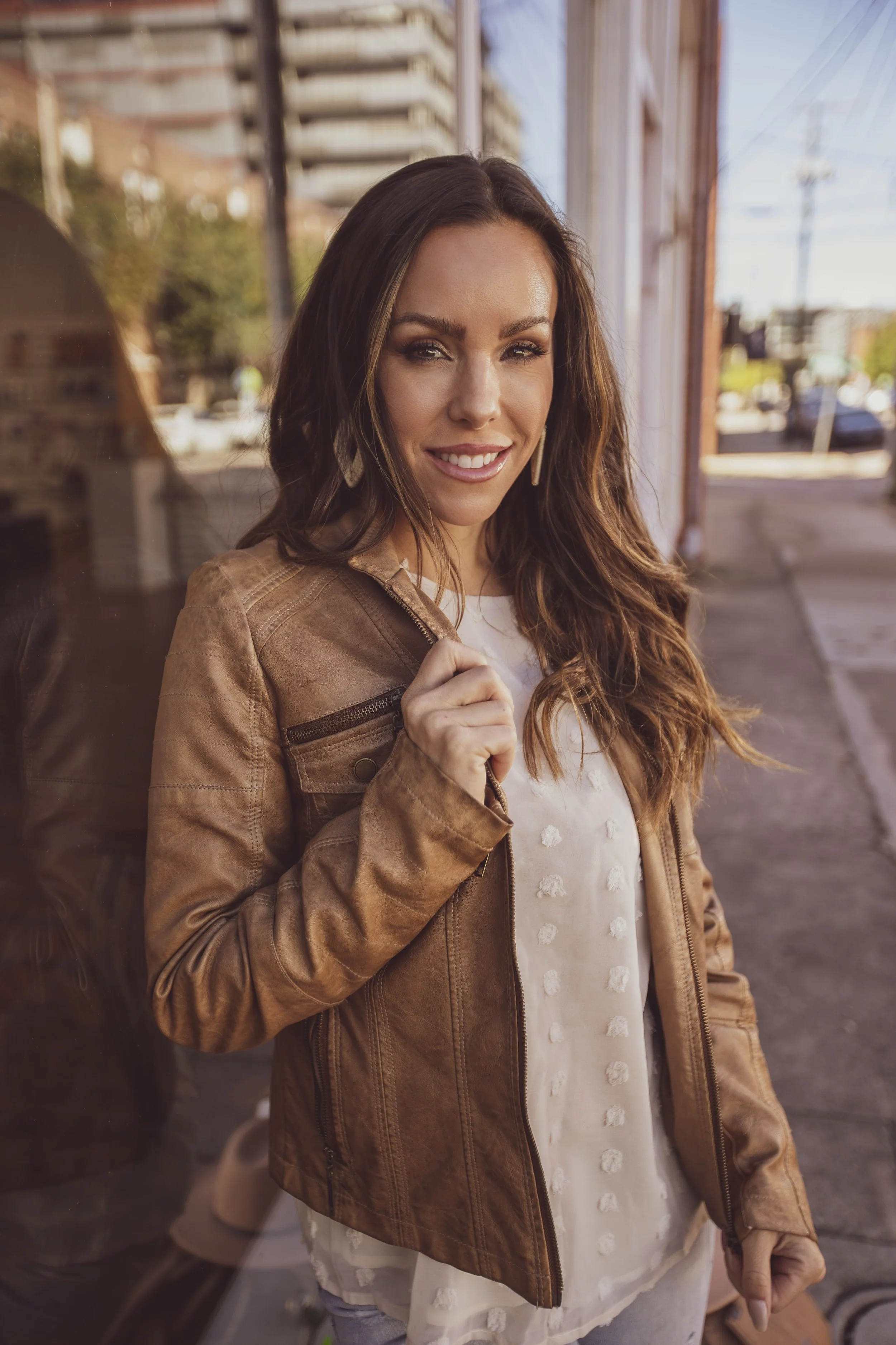 A woman with long brown hair, wearing a tan leather jacket over a white top, standing outdoors on a city sidewalk, smiling at the camera. BoldVision Studios