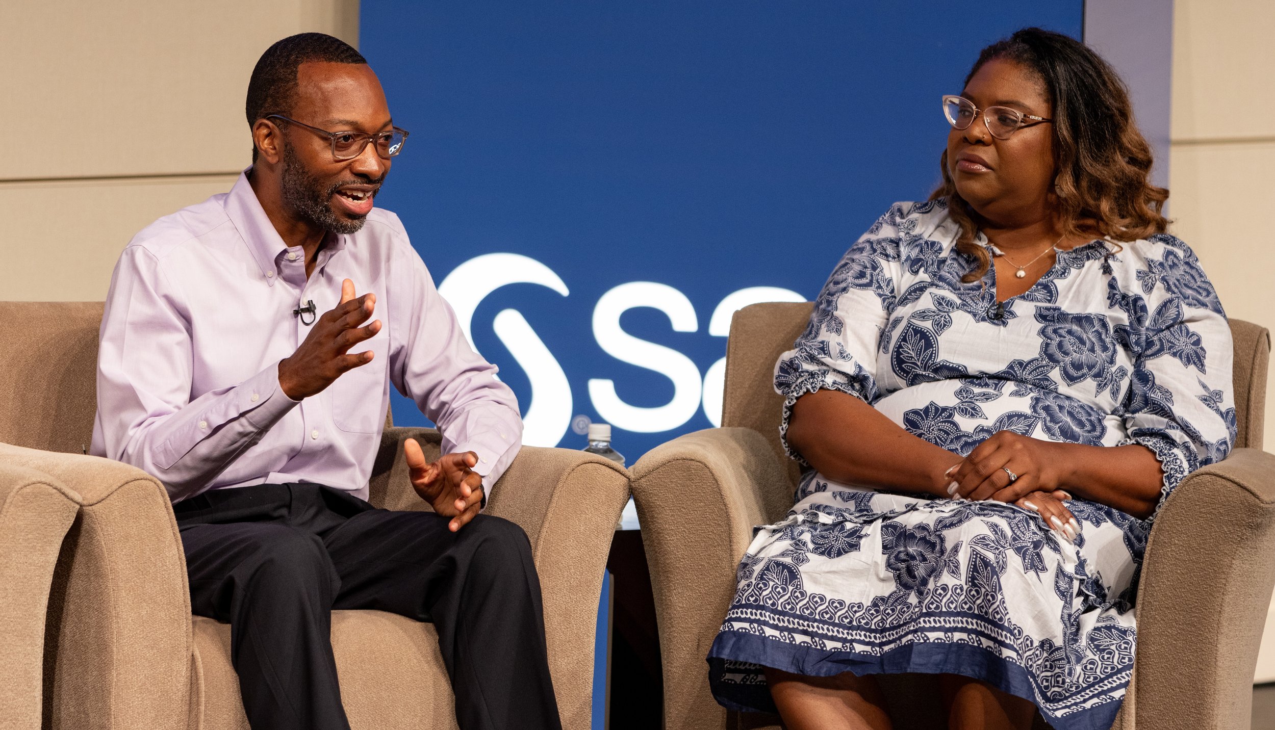 Two people sitting on beige armchairs having a discussion in front of a blue background with a logo. BoldVision Studios