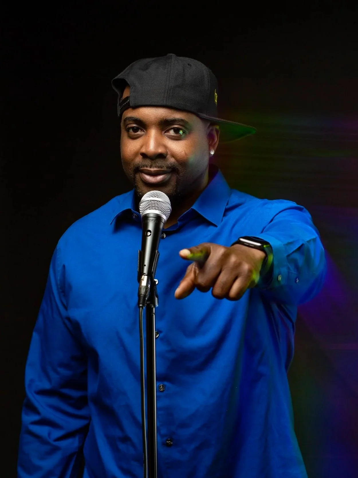 Man in a blue shirt wearing a black baseball cap, pointing towards the camera, standing in front of a microphone on a black background.