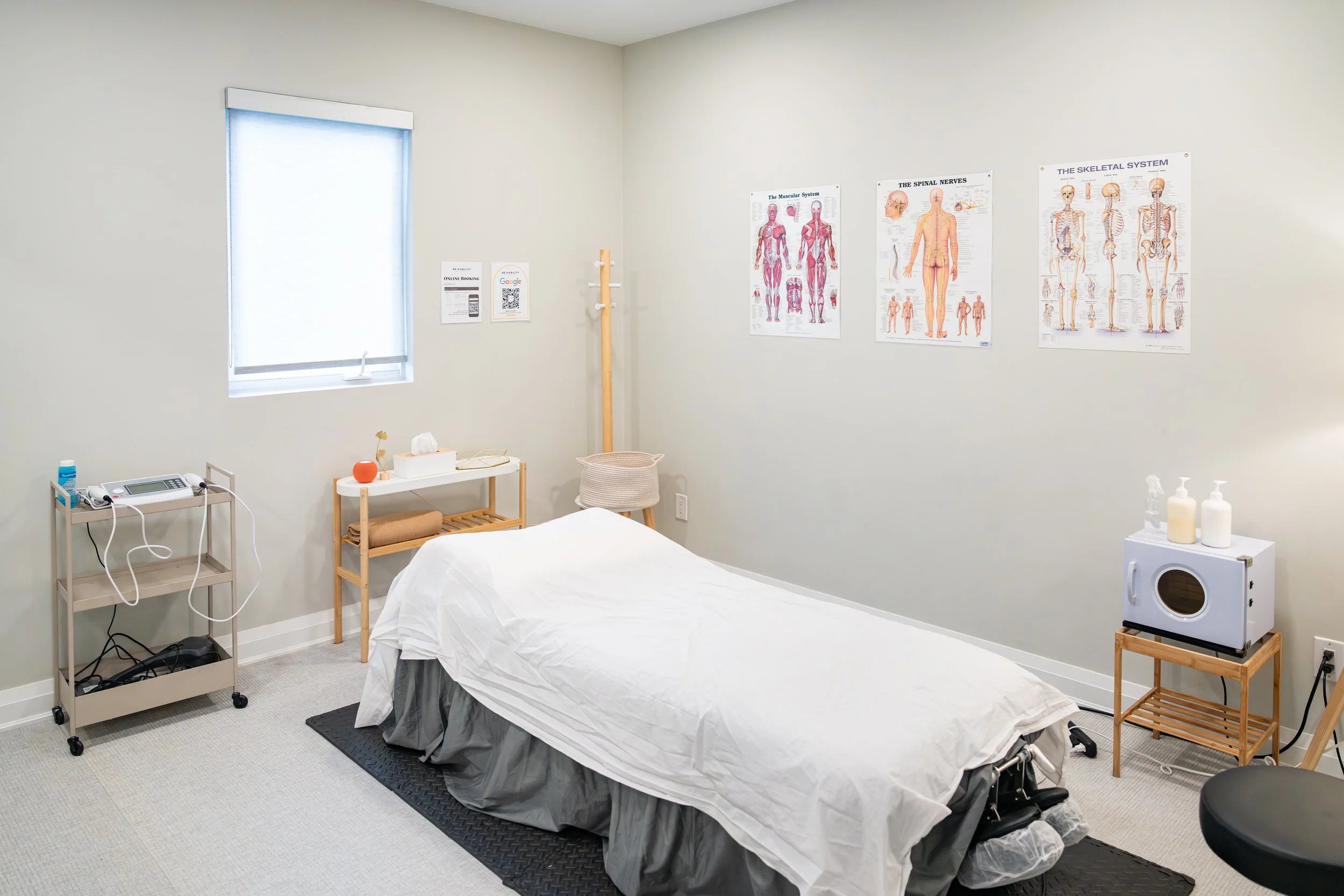A medical examination room with a bed covered in a white sheet, a window with blinds, anatomical posters on the walls, and medical equipment on tables and stands.