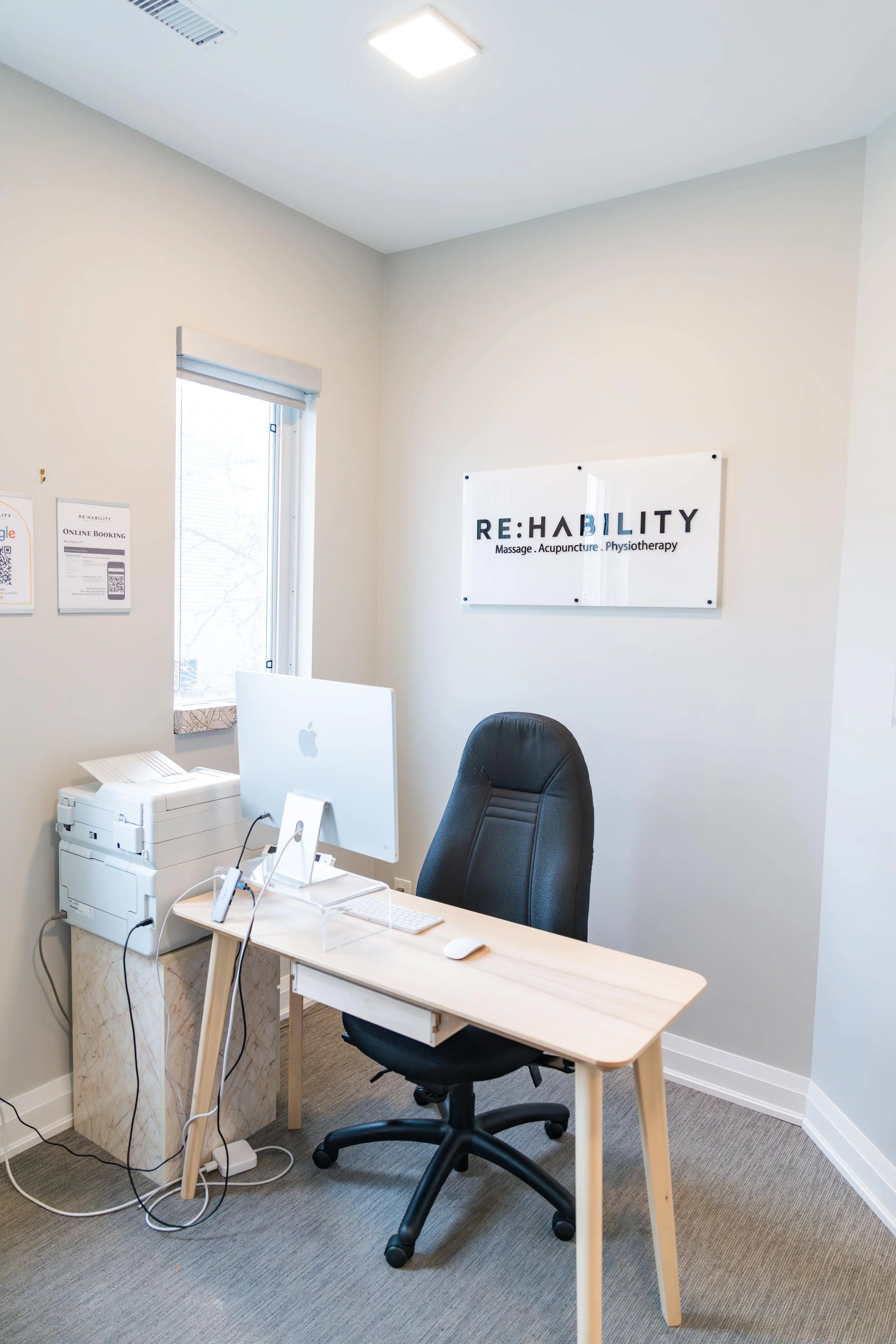 Office reception area with a wooden desk, black office chair, an Apple computer, a printer, and a sign on the wall that reads 'RE:HABILTY Massage, Acupuncture, Physiotherapy'.