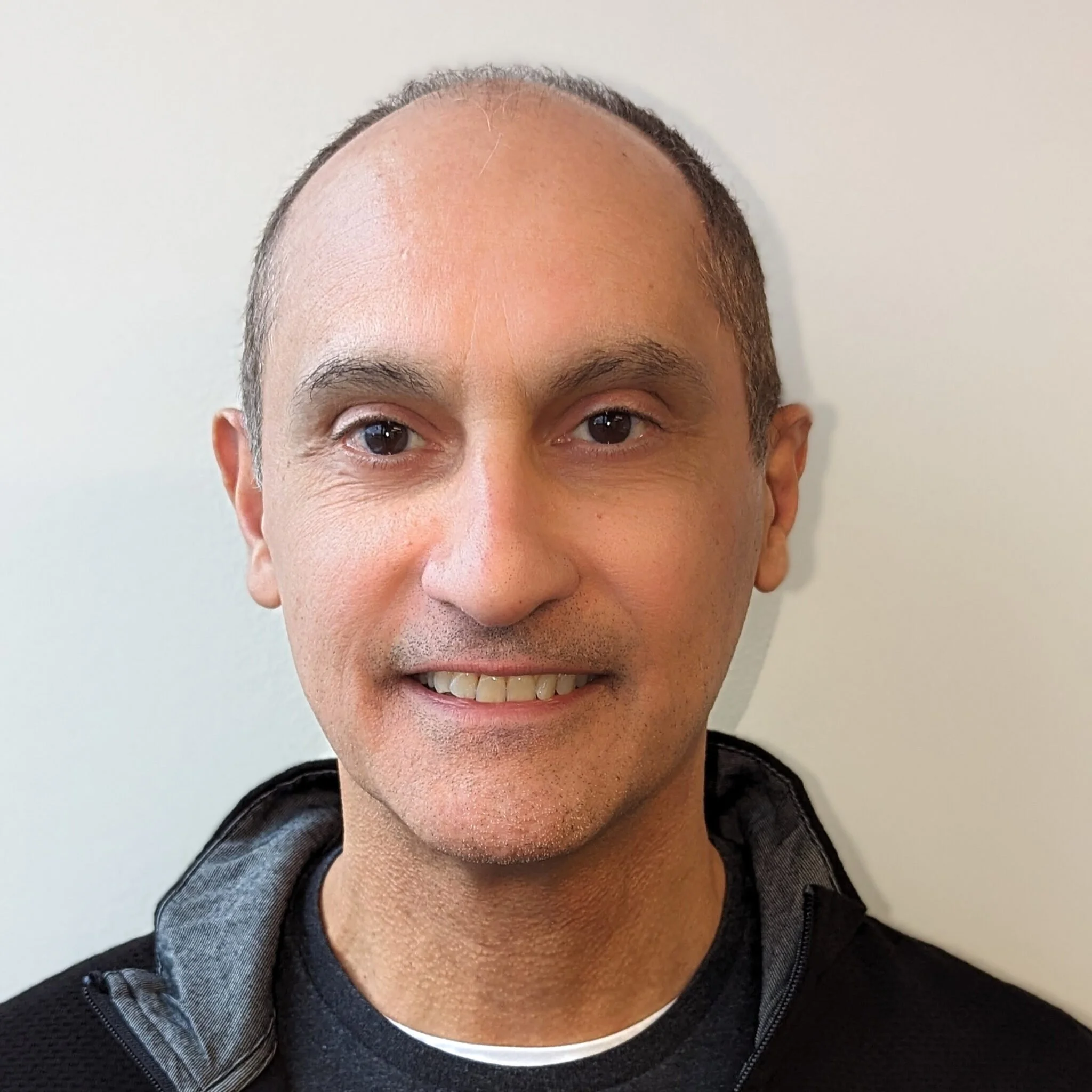 A smiling man with a shaved head and wearing a black jacket, standing in front of a plain light-colored wall.