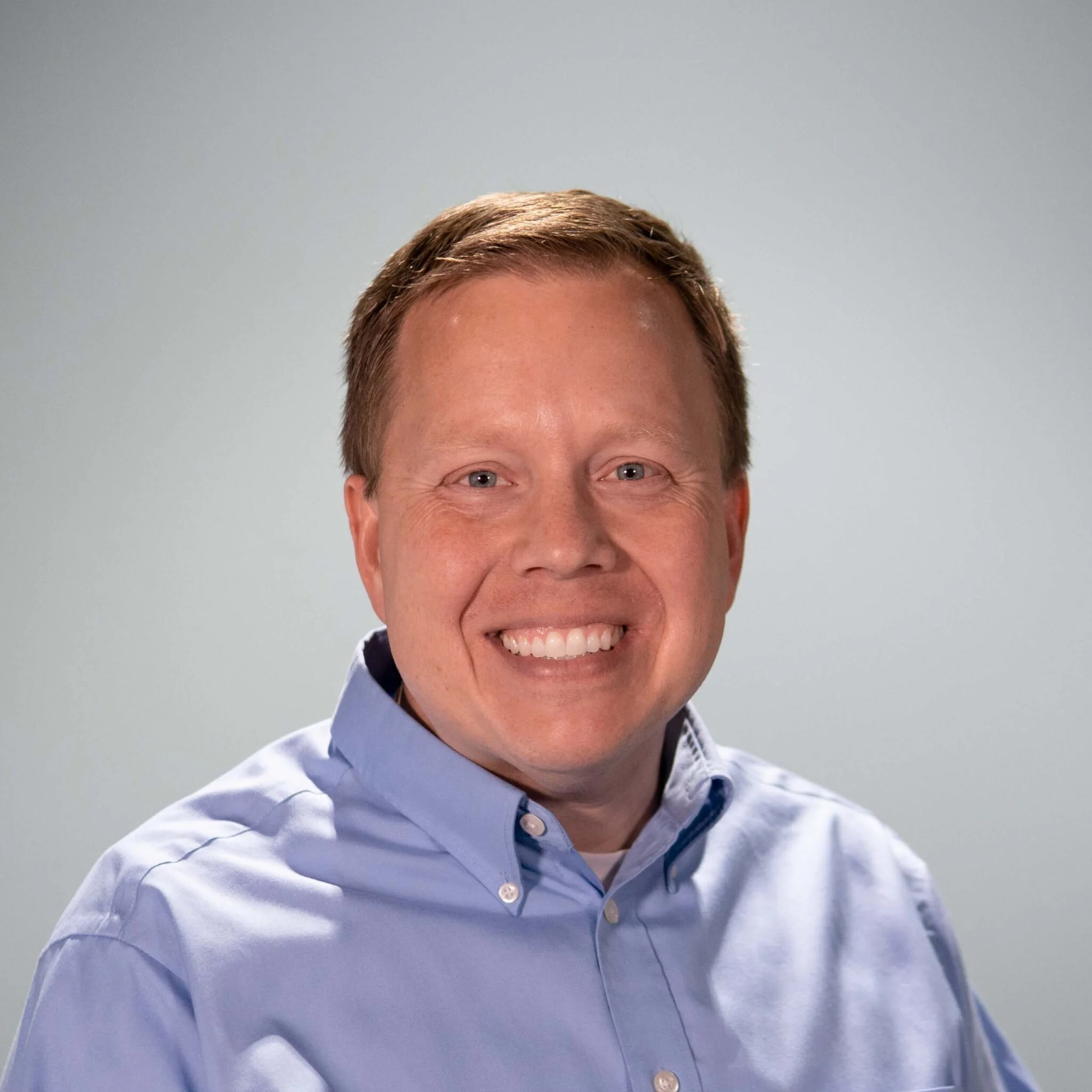 Close-up of a smiling man with blue eyes and short brown hair, wearing a light blue dress shirt against a plain light gray background.