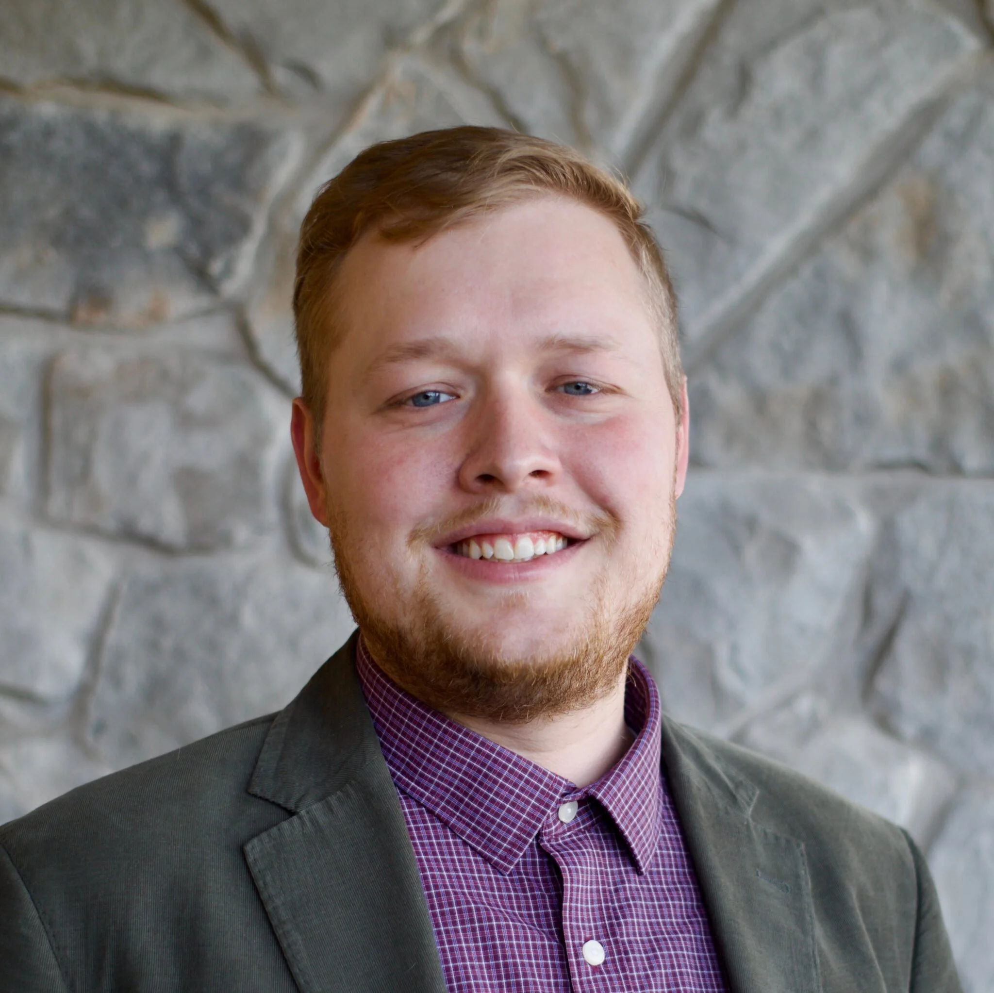 Close-up of a young man with red hair and blue eyes, wearing a dark blazer, purple checkered shirt, and carrying a backpack, standing against a stone wall wall.