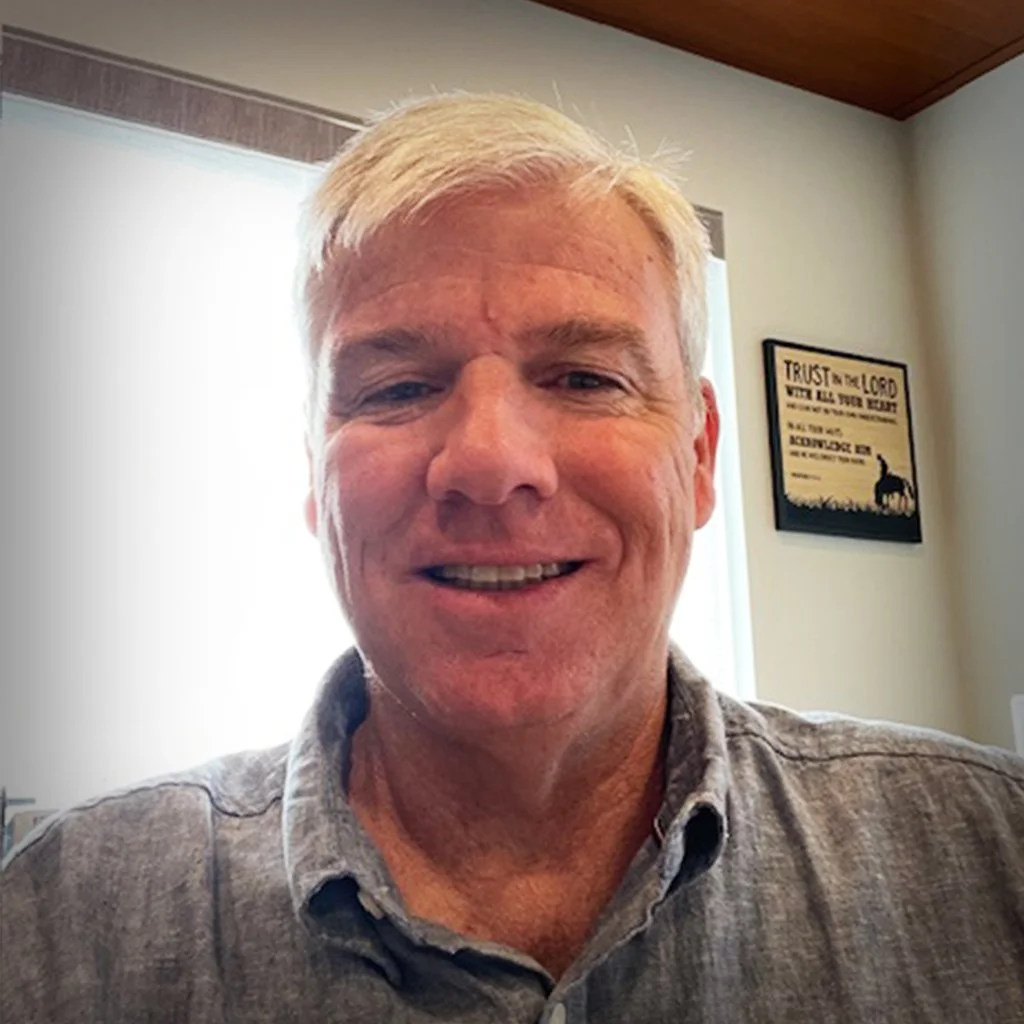 A smiling middle-aged man with short, light-colored hair taking a selfie indoors. There is a window and a framed picture on the wall behind him.