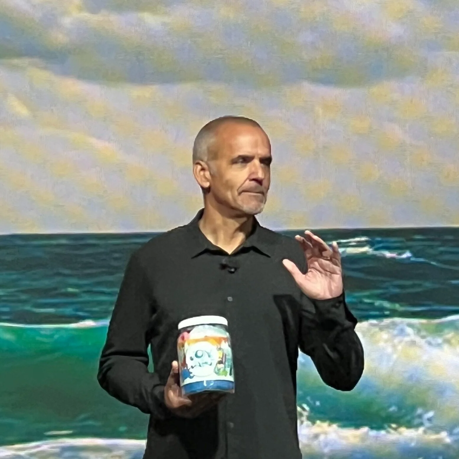 A man with a shaved head and dark shirt holding a colorful object in front of a backdrop of ocean waves and a cloudy sky.