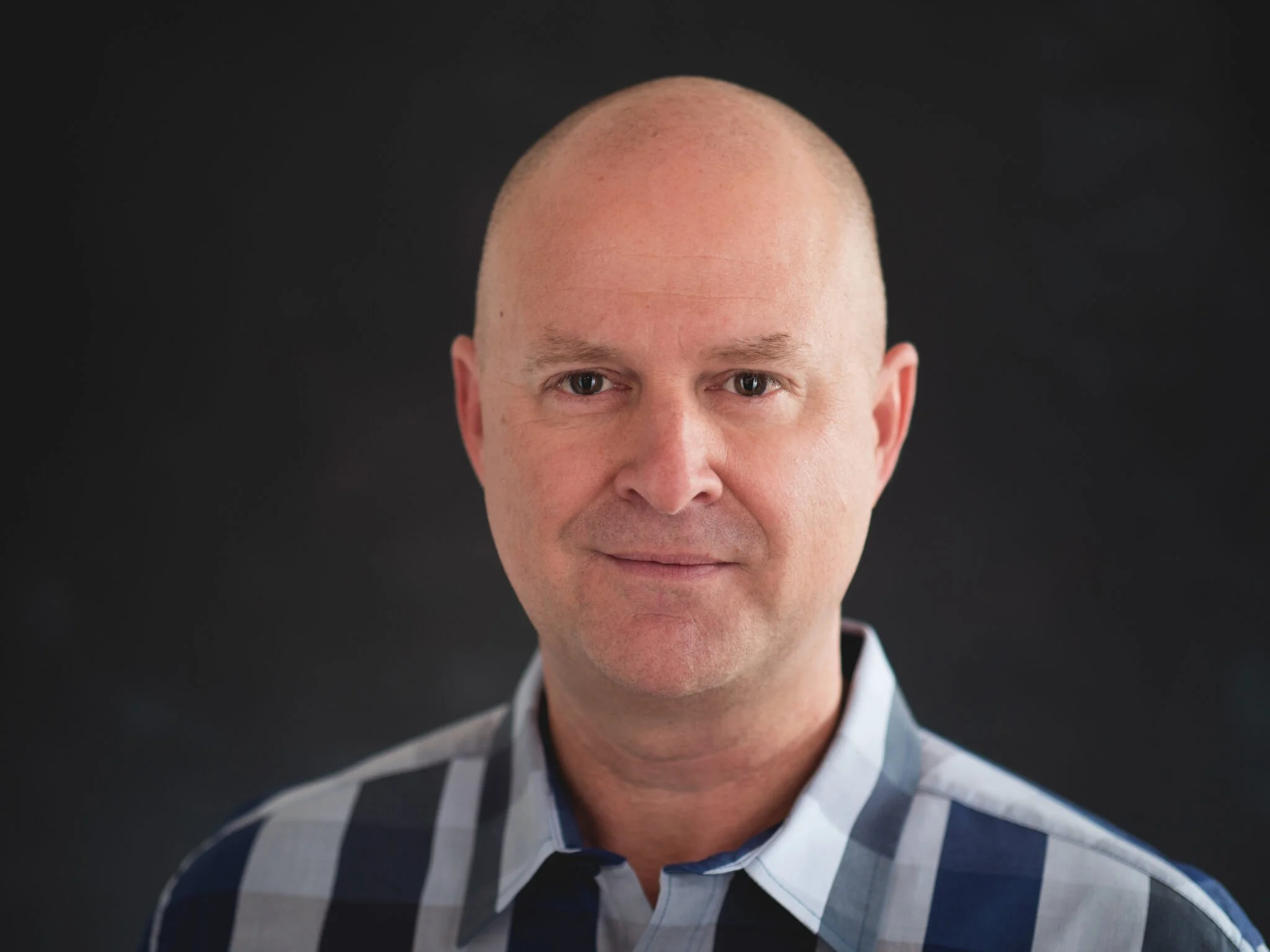 A close-up portrait of a man with a bald head, wearing a dark-colored, plaid shirt with shades of gray and white, against a plain black background.