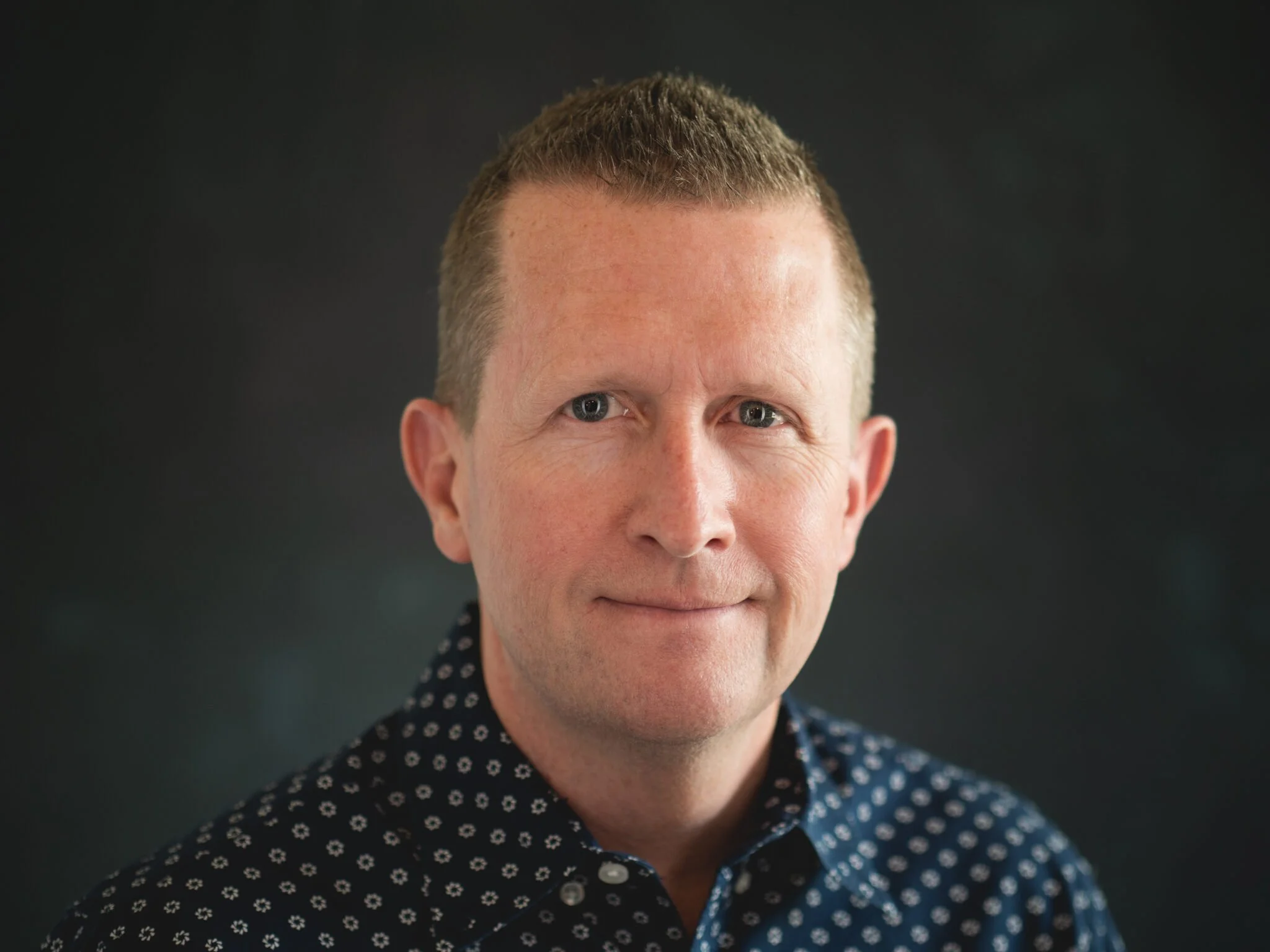 Close-up of a man's face, showing short brown hair, blue eyes, and wearing a dark shirt with a small pale pattern, against a dark background.