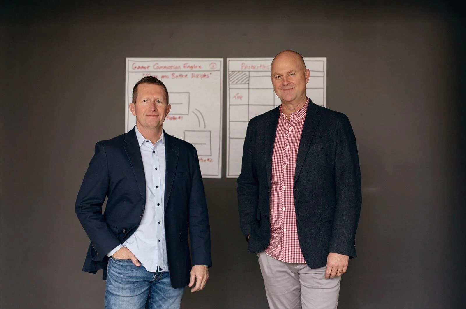 Two men in business casual attire standing in front of a dark wall with two large charts on paper behind them, one labeled 'Great Conversion Engine' and the other 'Prioritization'.
