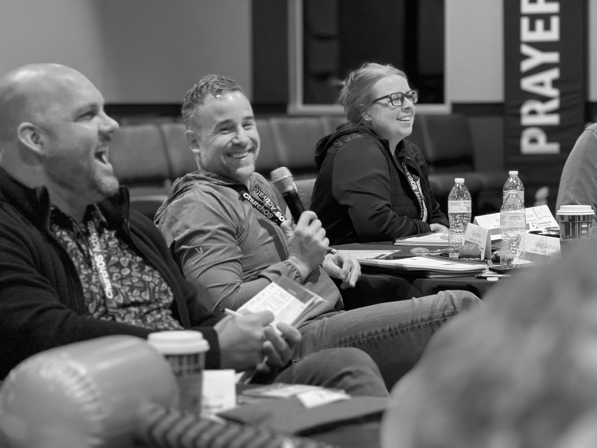 Three people sitting at a conference table, smiling and engaging, with water bottles and papers in front of them, in a meeting or discussion setting.