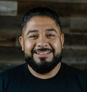 A man with short dark hair, a beard, and a mustache smiling, wearing a black shirt against a wooden background.