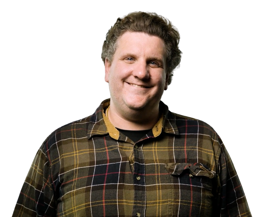 A smiling man with light brown, curly hair wearing a brown plaid shirt with a yellow collar.