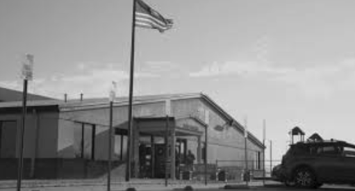 A black and white photo of a building with a flagpole flying an American flag, a pickup truck parked in front, and a fountain in the foreground.