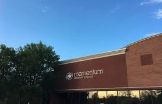 Exterior of a brick building with a sign that reads 'Momentum' and 'Wellness Center' underneath it, with trees and a blue sky in the background.
