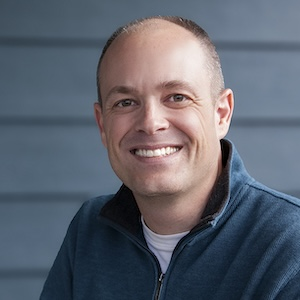 A man with a receding hairline smiling, wearing a blue jacket over a white shirt, in front of a blue wooden background.