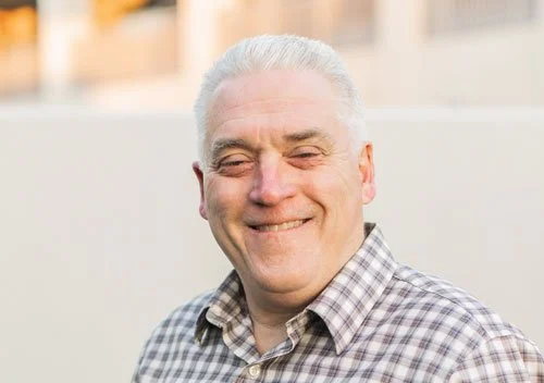Smiling older man with gray hair wearing a checkered shirt outdoors.