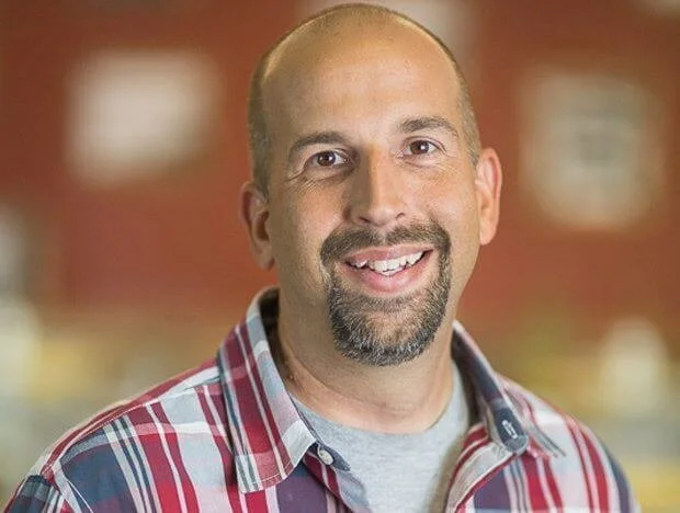 A smiling man with a goatee and bald head, wearing a plaid shirt, standing indoors with a blurred background.