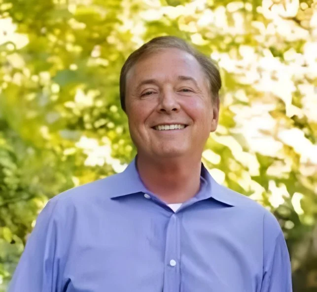 A smiling man in a blue collared shirt outdoors with blurred green foliage in the background.