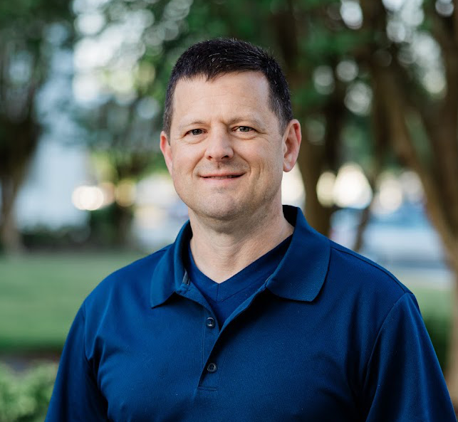 Portrait of a man with short dark hair, wearing a blue collared shirt, standing outdoors with blurred trees in the background.