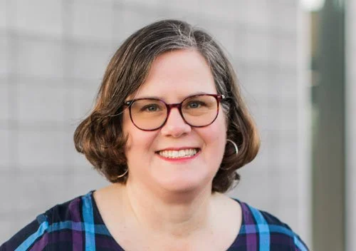 A woman with shoulder-length brown hair, glasses, and hoop earrings, smiling in front of a gray textured background.