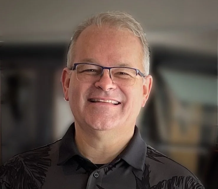 A smiling middle-aged man with glasses, wearing a black collared shirt, standing indoors.