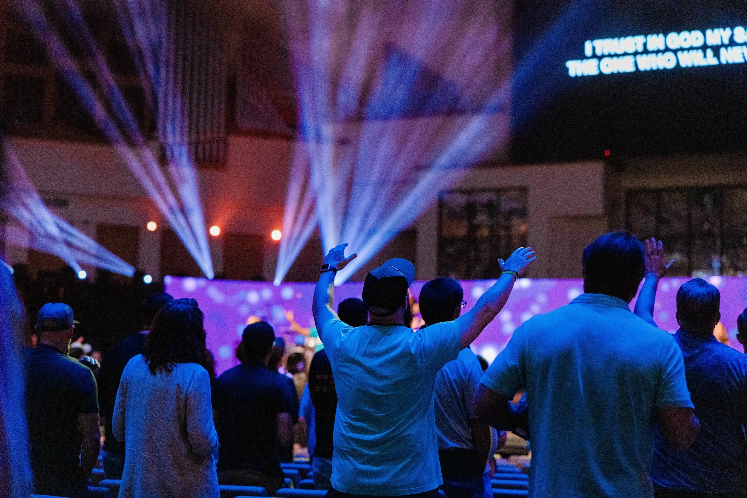 Crowd of people at an indoor event or concert, facing a stage with blue and purple lighting effects, with some raising their hands. There is a large screen or projection displaying the text 'I TRUST IN GOD MY LORD THE ONE WHO WILL NEVER'.