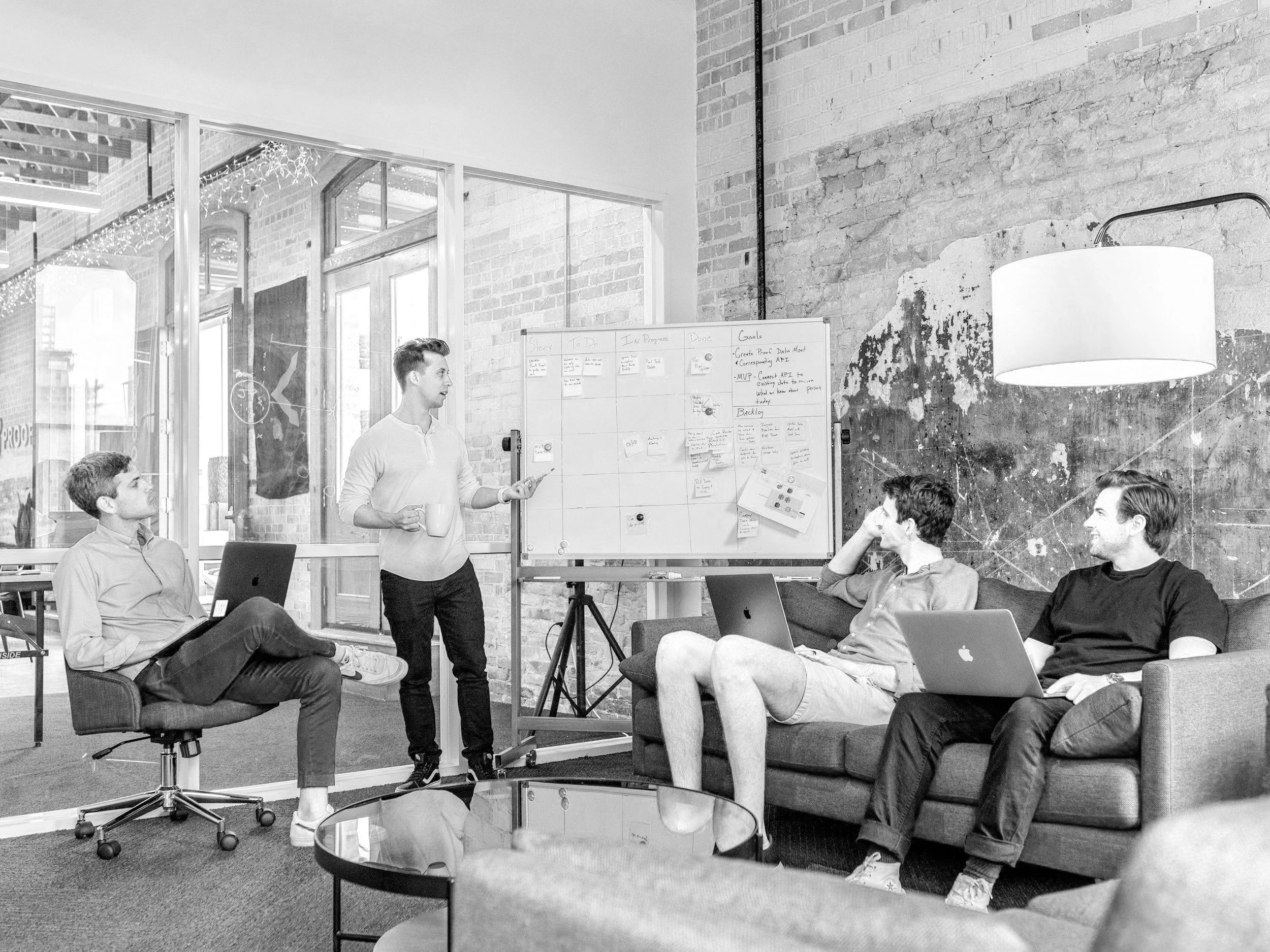 A group of young professionals in a modern office with exposed brick walls, participating in a team meeting. One person is standing in front of a whiteboard, presenting, while others sit on couches and chairs with laptops and notes, listening and engaging.